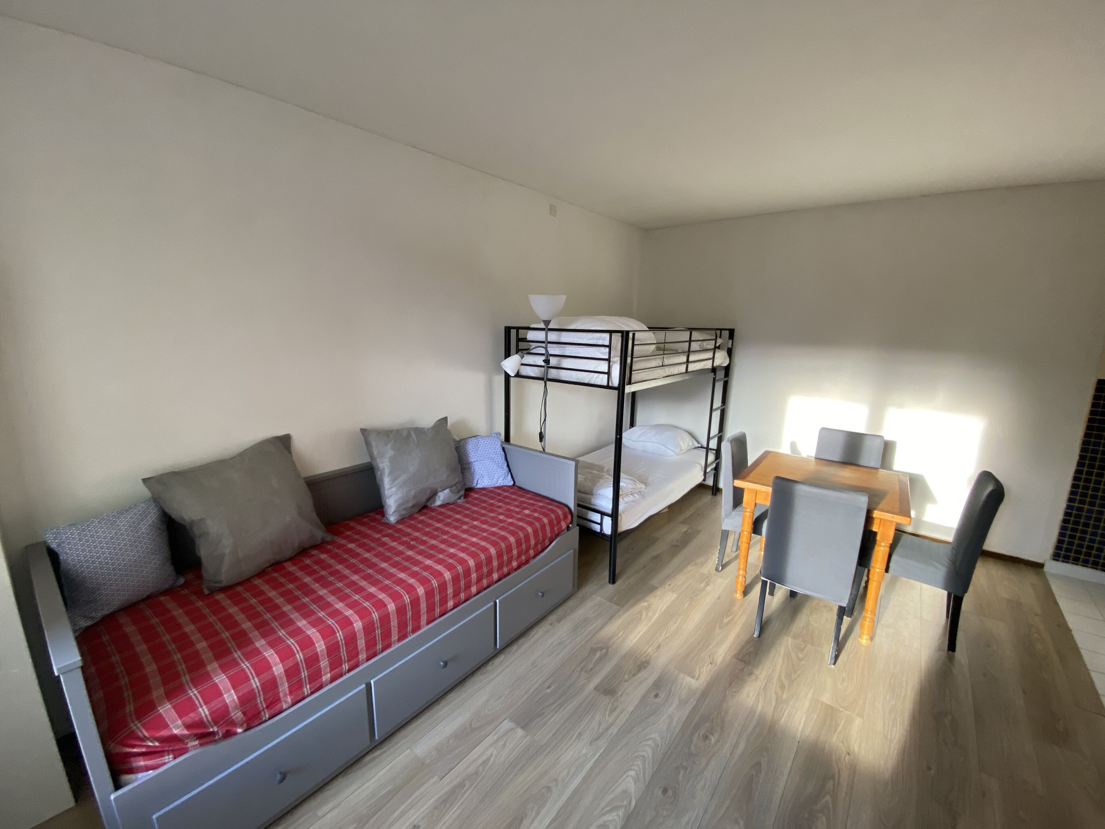 A dormitory room with a bunk bed, a sofa bed, a small table with chairs, and a lamp.