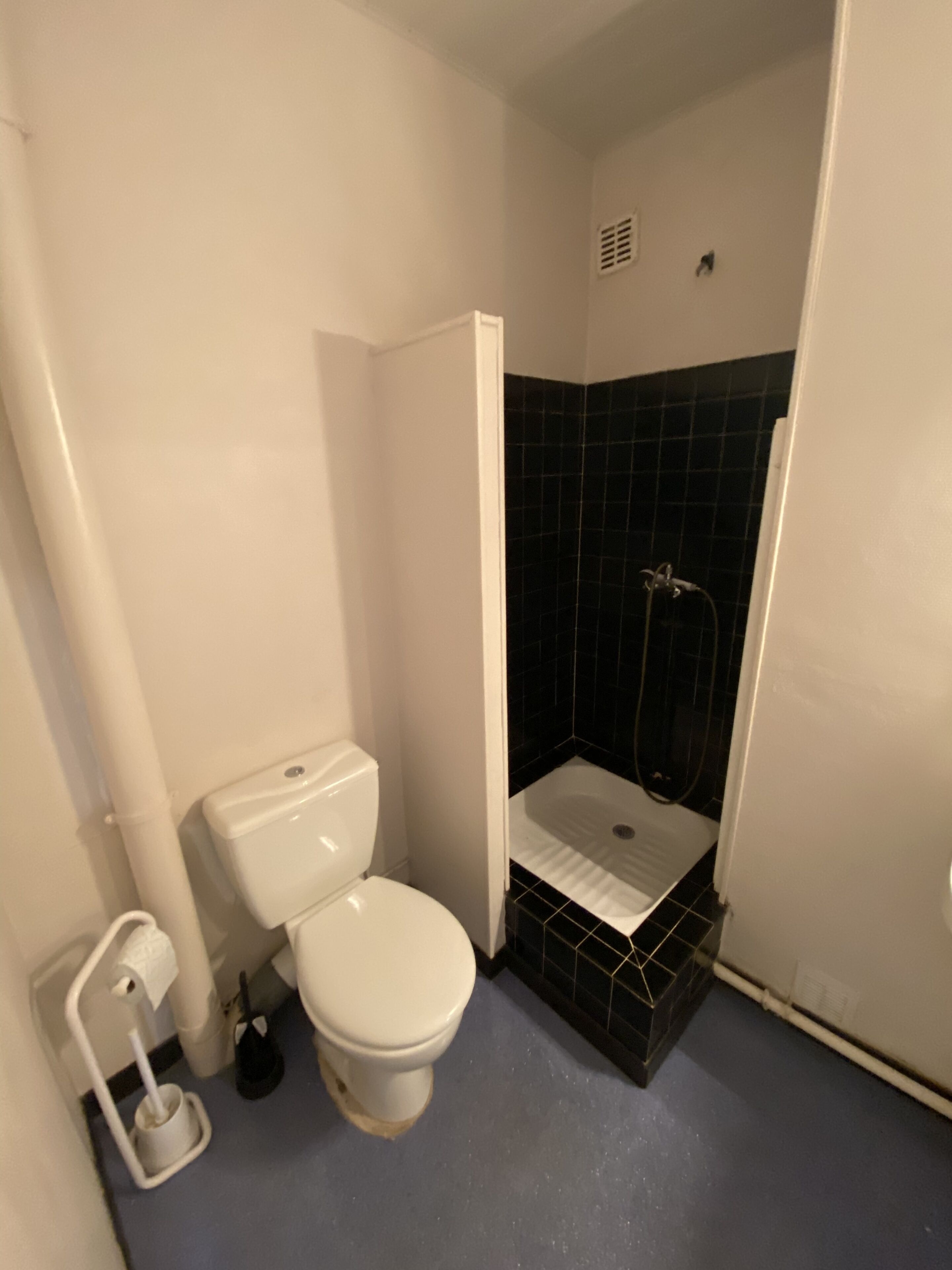 Une salle de bain avec des toilettes blanches, un espace douche avec des carreaux noirs et un sol bleu.