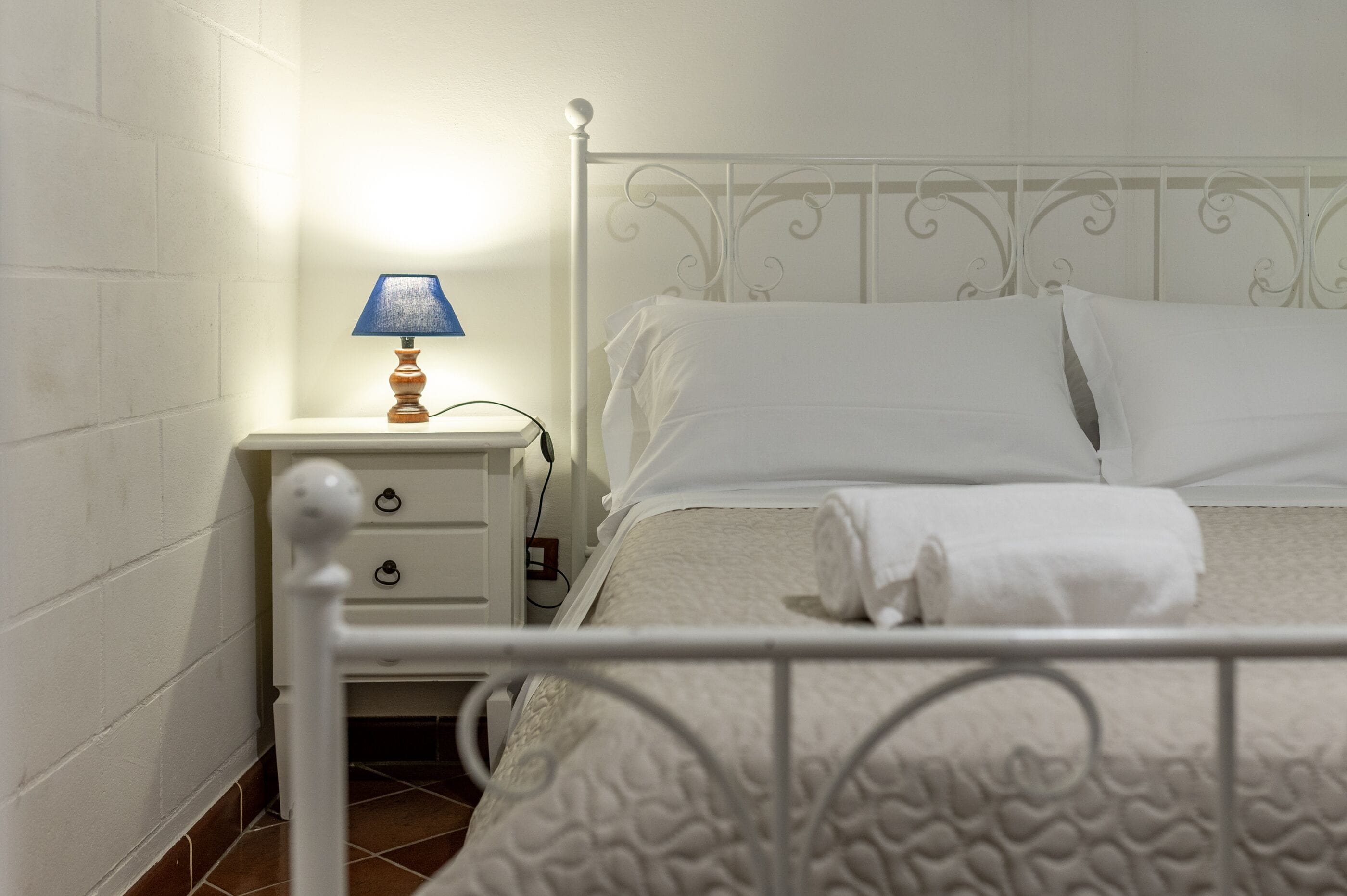 A neatly made bed with white linens, a bedside table with a lamp, and a white metal headboard.
