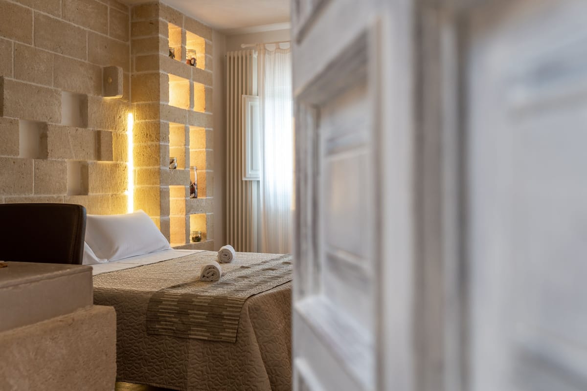 A hotel room with a stone wall, a bed with a textured blanket, and a small table with a chair.
