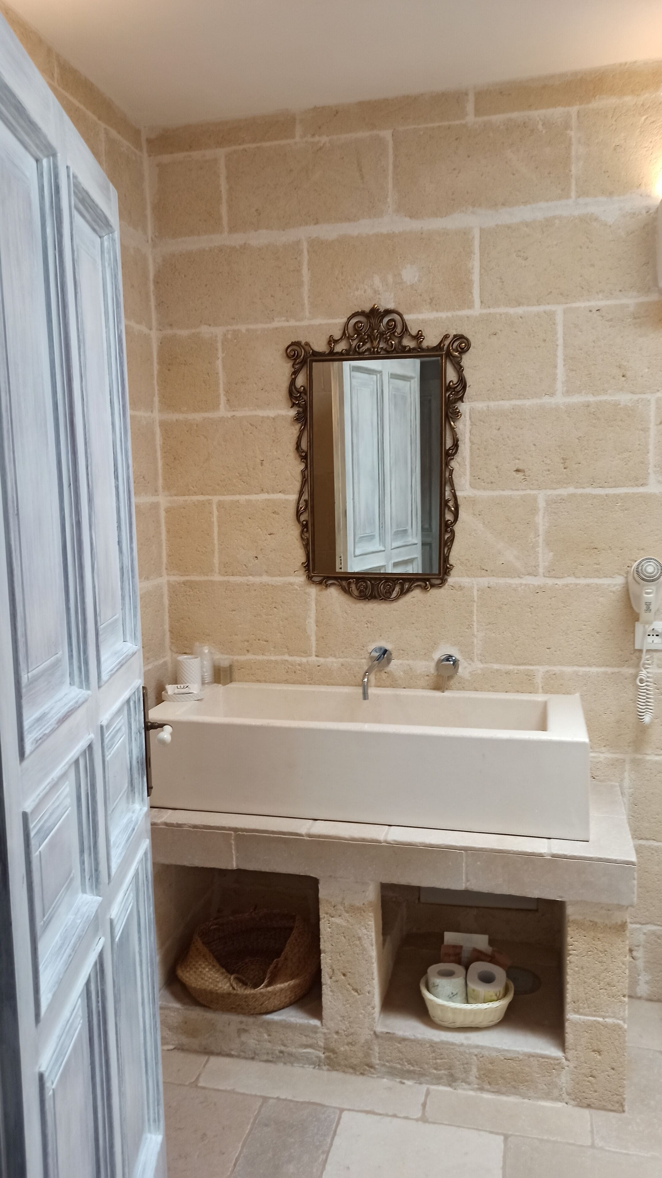 Une salle de bain avec un lavabo blanc, un miroir doté d’un cadre orné et un sèche-cheveux fixé au mur.