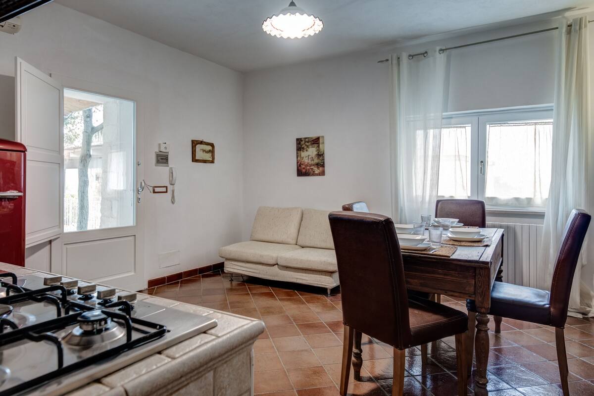 A kitchen with a gas stove, a dining table with chairs, a sofa, and a window with curtains.