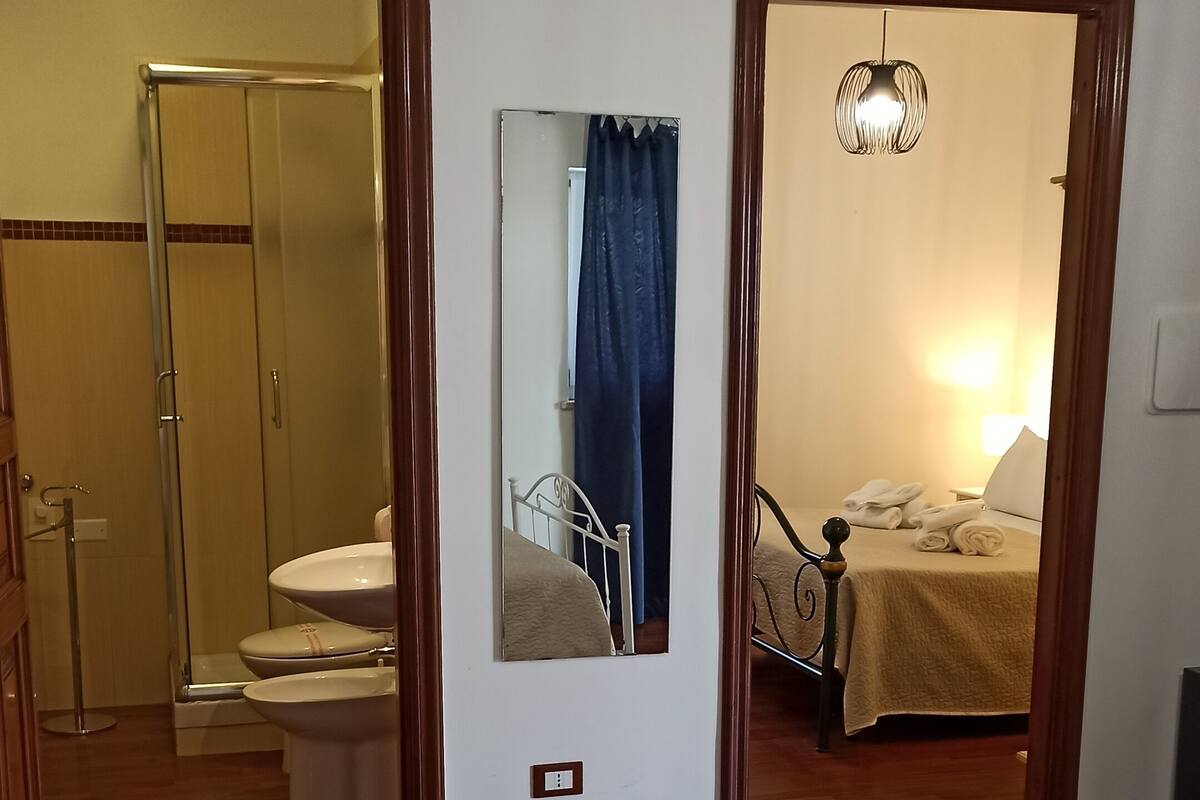 A hotel room with a bed, a bathroom with a toilet and sink, a mirror, and a bedside table with towels.