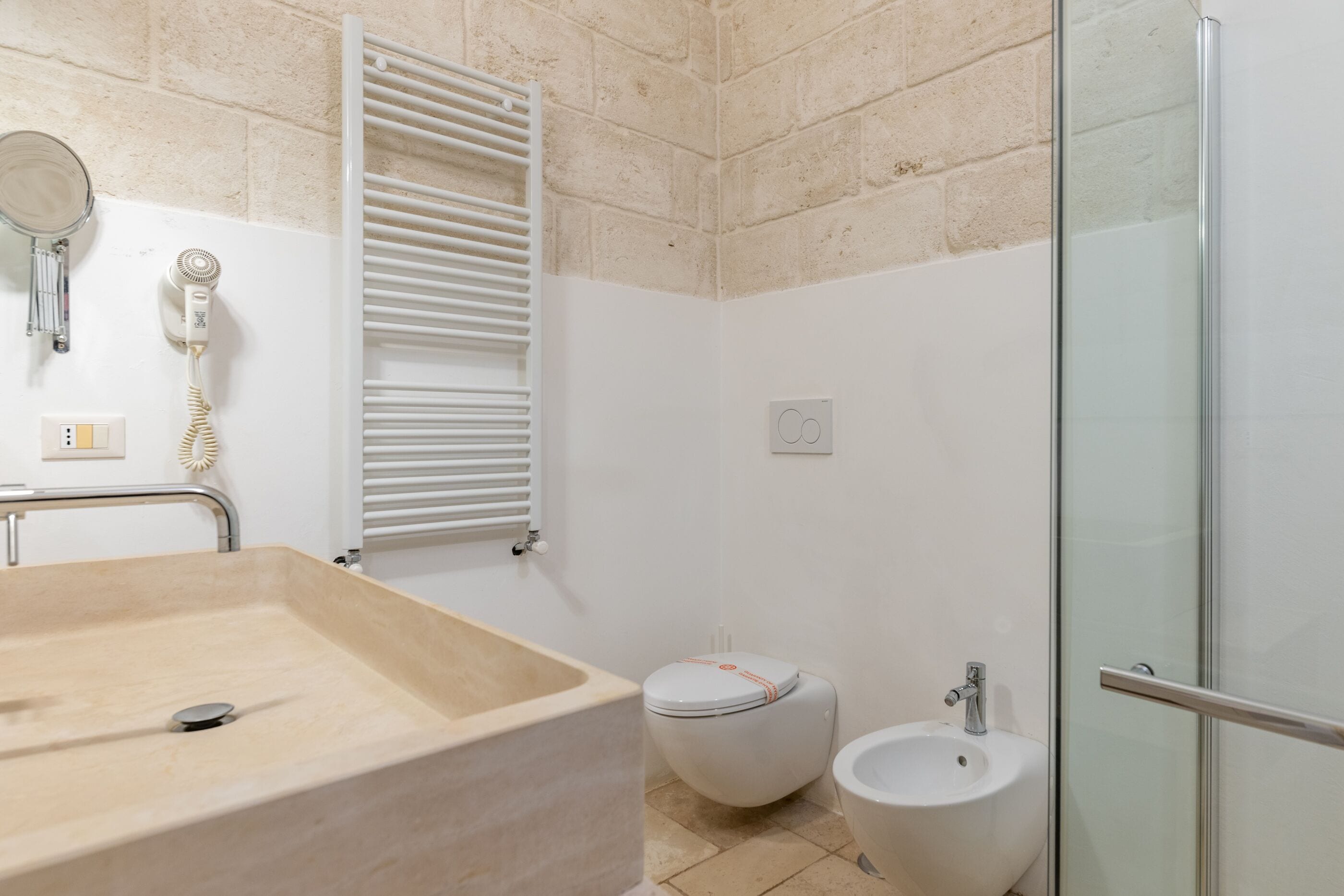 A bathroom with a beige sink, a toilet with a white lid, a bidet, and a glass shower enclosure.