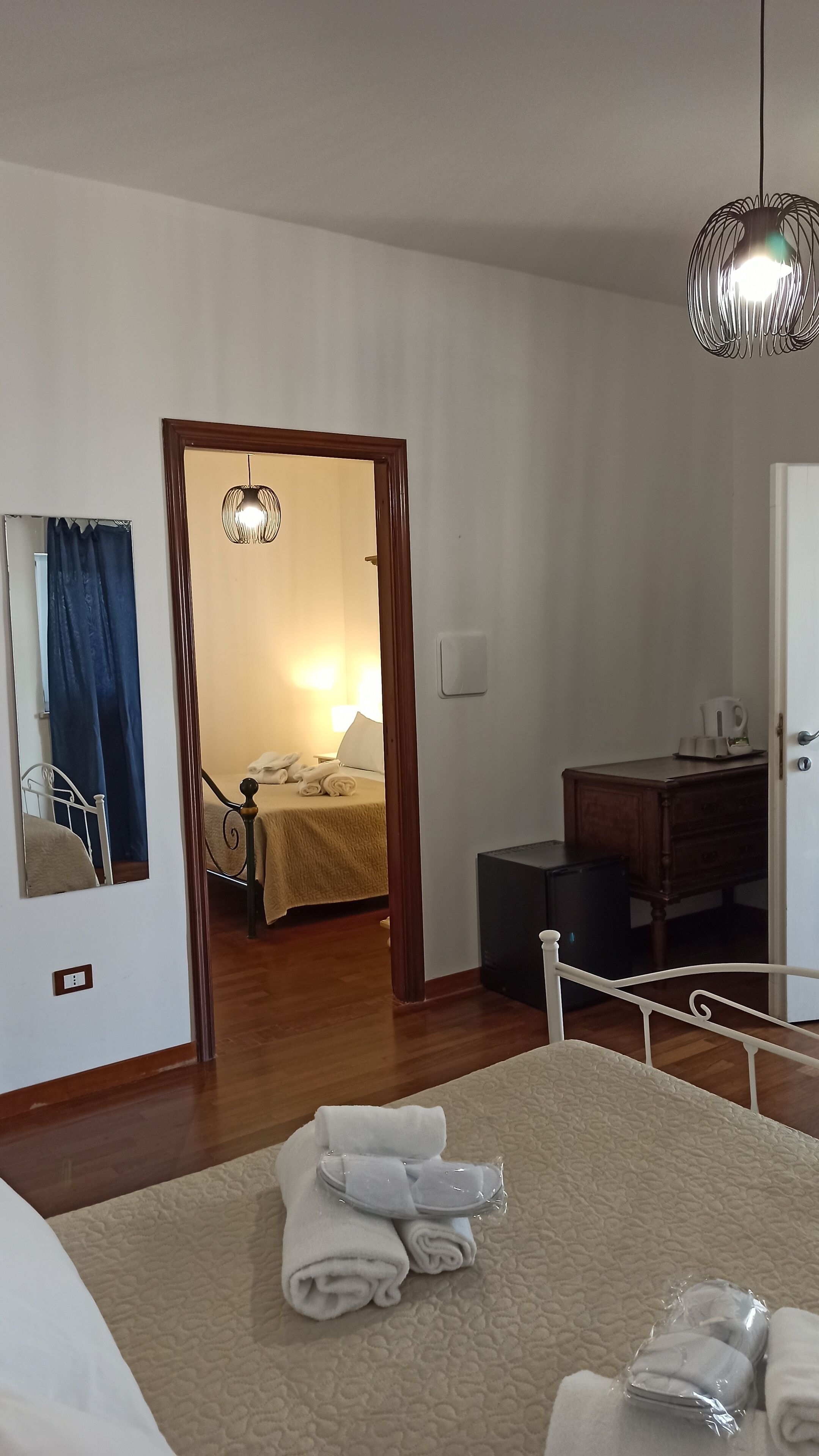 Quarto de hotel com cama, escrivaninha, cadeira, espelho, porta e luminária pendente.