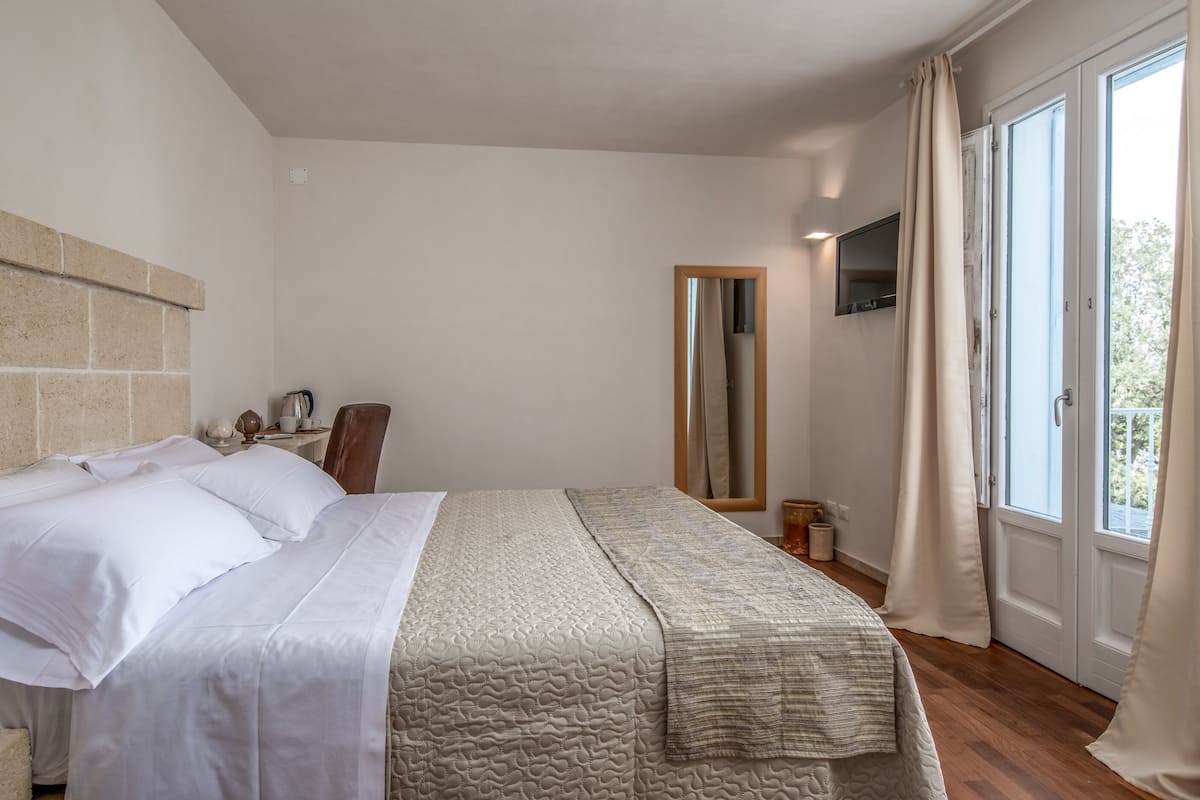 A bedroom with a large bed, a wooden headboard, a mirror, a bedside table with a lamp, and a window with curtains.