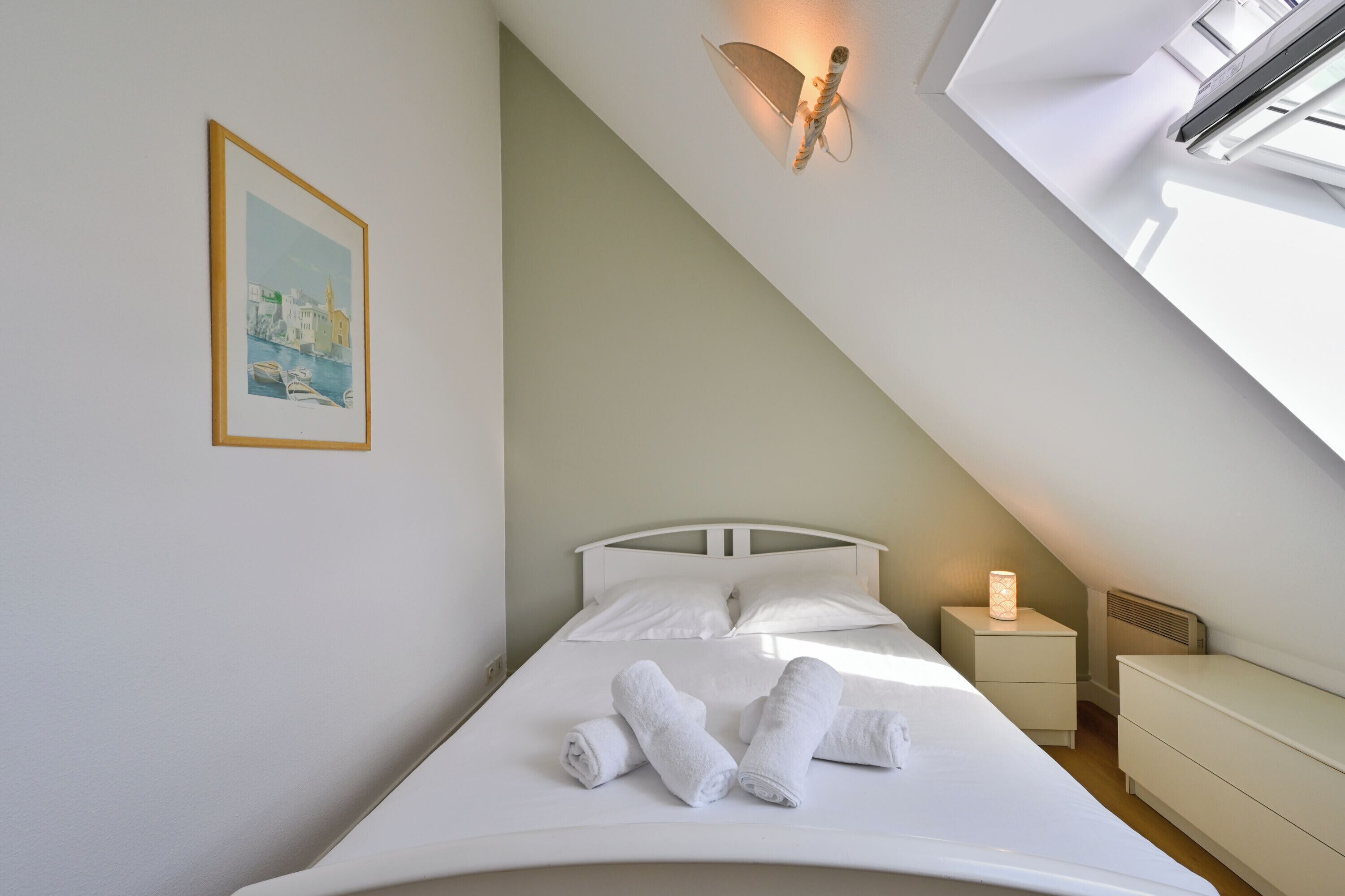 Quarto com cama, almofadas, mesa de cabeceira, abajur, quadro emoldurado e teto inclinado.