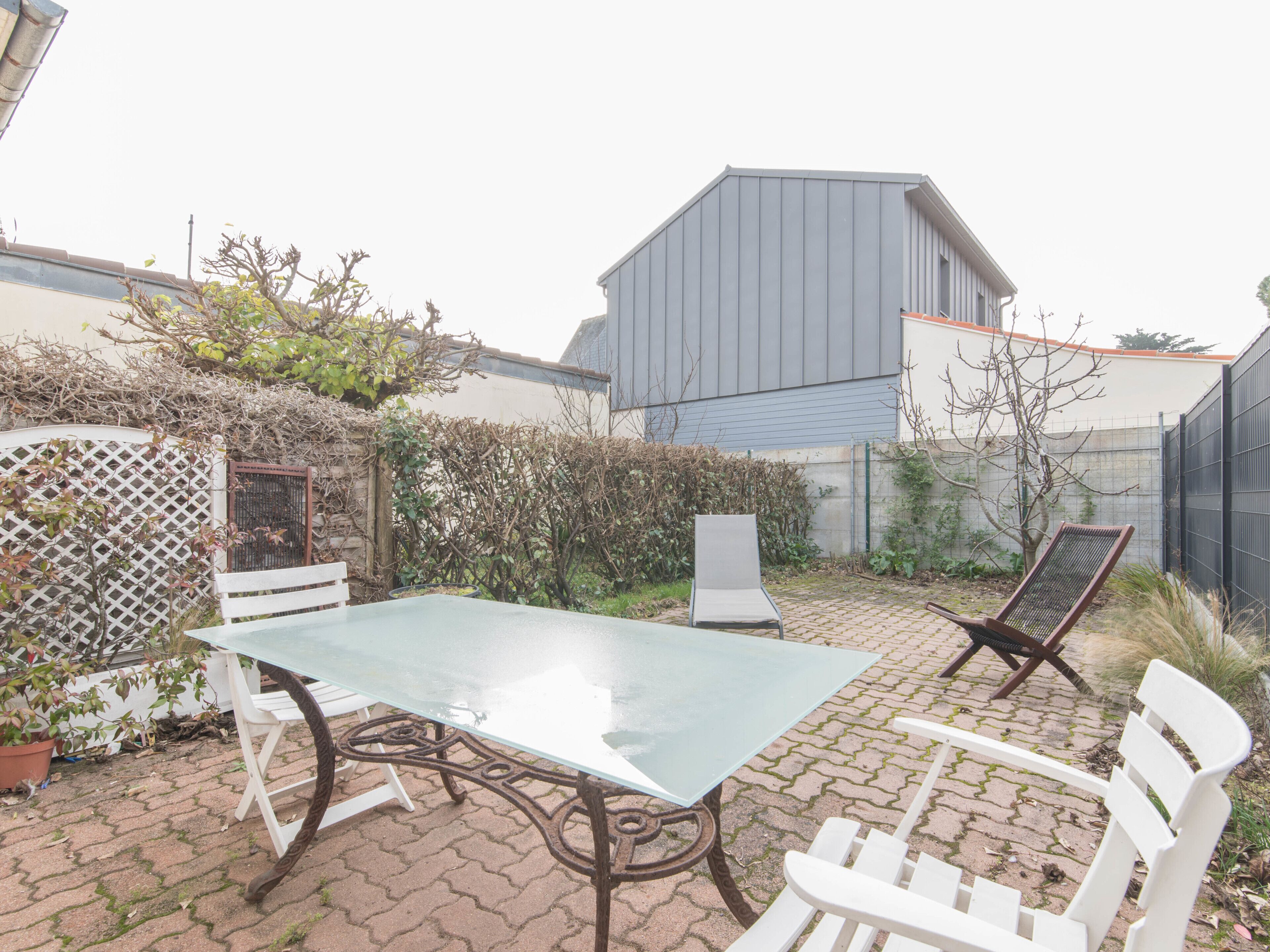 A patio with a glass table and white chairs, surrounded by a brick wall and a hedge.