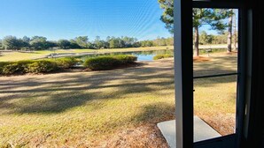 Une vue à travers une porte vitrée sur un terrain de golf, avec un étang, des arbres et un ciel dégagé.
