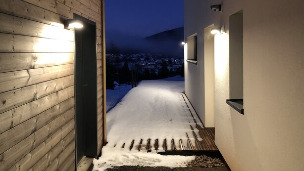 Camino nevado que conduce a un edificio con revestimiento de madera e iluminación exterior.