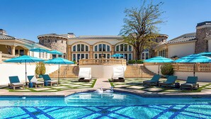 A swimming pool with a geometric tile pattern, surrounded by lounge chairs and umbrellas, with a building featuring arched windows and a stone facade in the background. - Chateau Dumont - 15 bedroom mansion (Rancho Mirage)