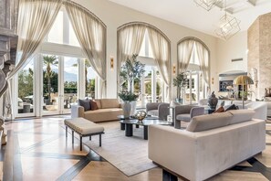A spacious living area with a large sofa, armchairs, a coffee table, and a dining table. There are large windows with curtains, a fireplace, and a view of the outdoors.