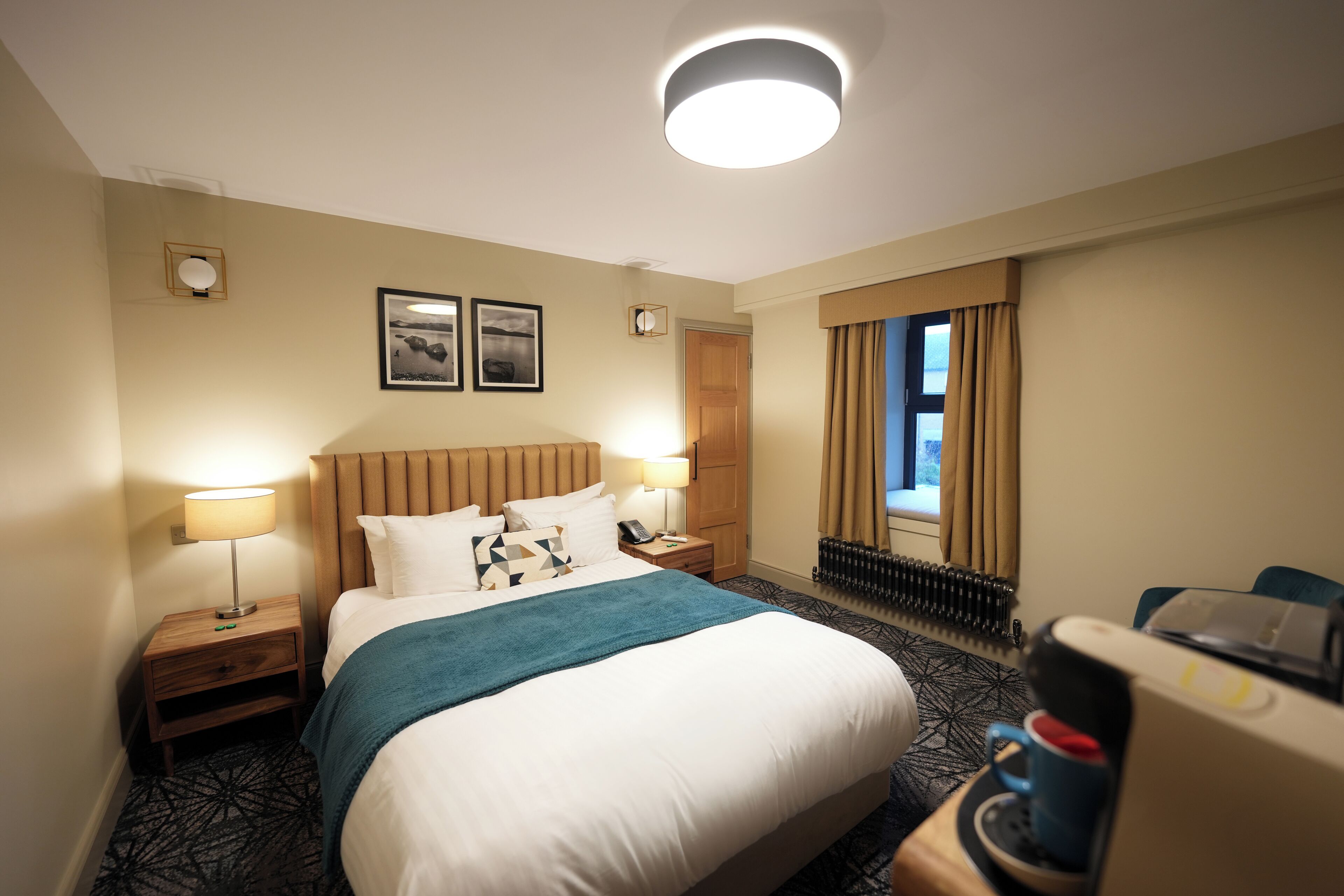 A hotel room with a large bed, two bedside lamps, a nightstand, a window with curtains, and a coffee machine.