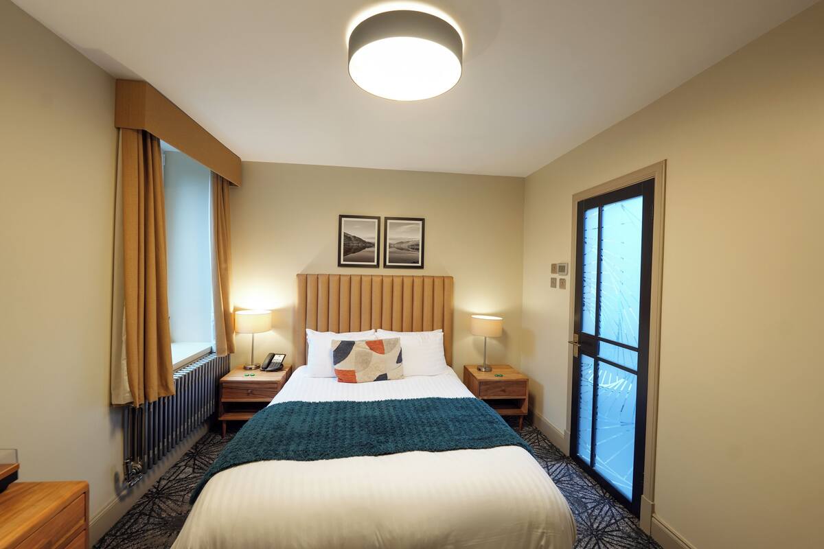 A hotel room with a bed, two bedside tables, a nightstand, a window with curtains, a radiator, and a door with frosted glass.