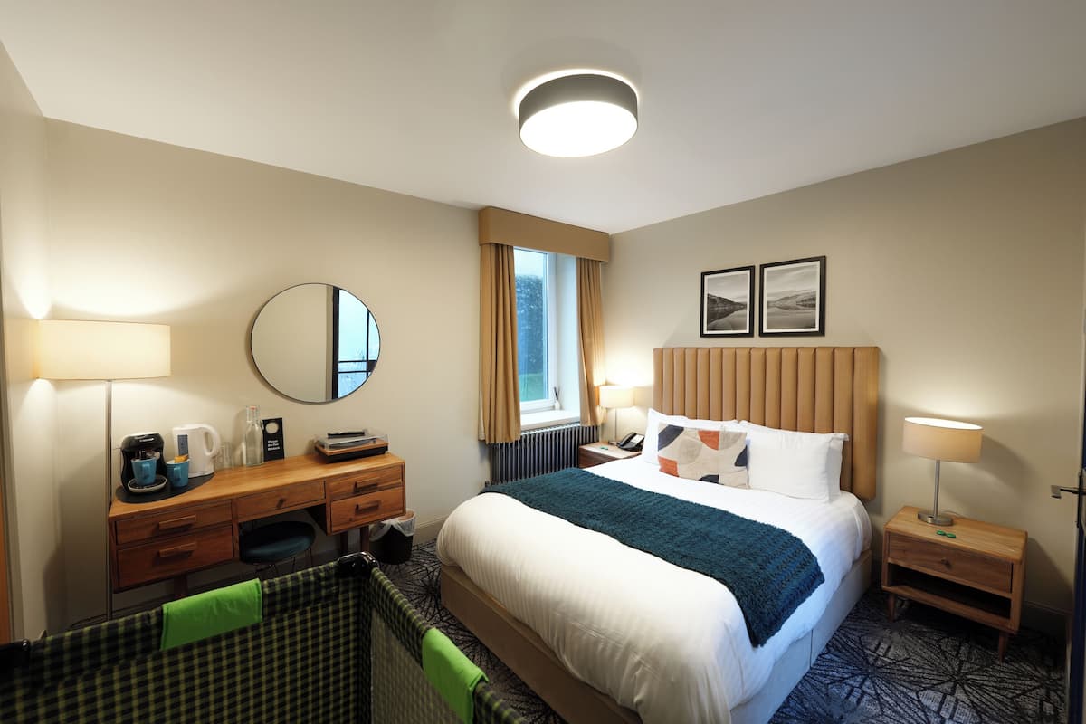 A hotel room with a bed, bedside tables, a desk, a chair, a mirror, a window with curtains, and a wall-mounted picture.