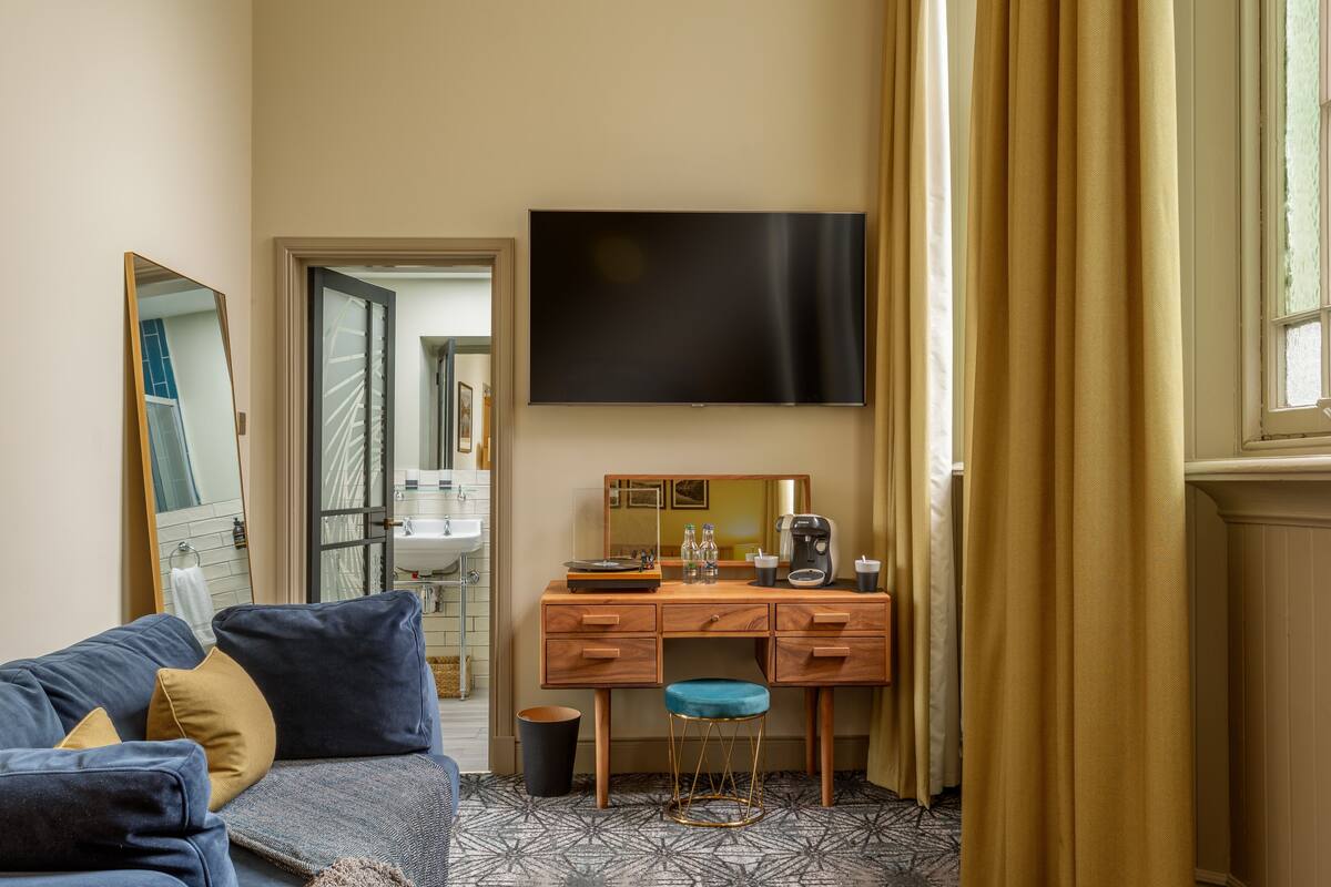 A hotel room with a sofa, a desk with a mirror, a flat-screen TV, and a window with curtains.
