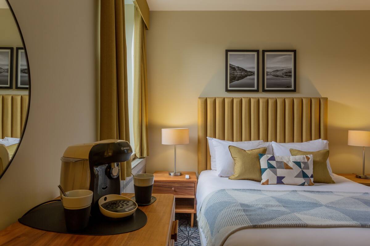 A hotel room with a bed, bedside lamps, a coffee maker, a cup, and a small table with a spoon.