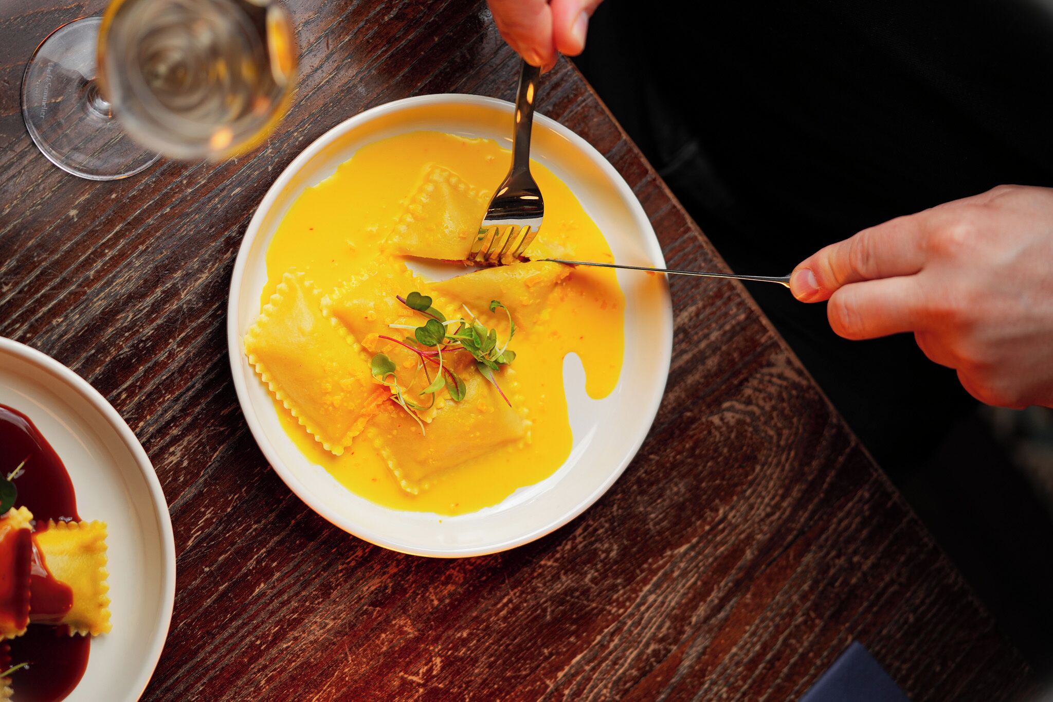 A plate of ravioli with sauce and garnish, a fork, and a glass of wine on a wooden table.