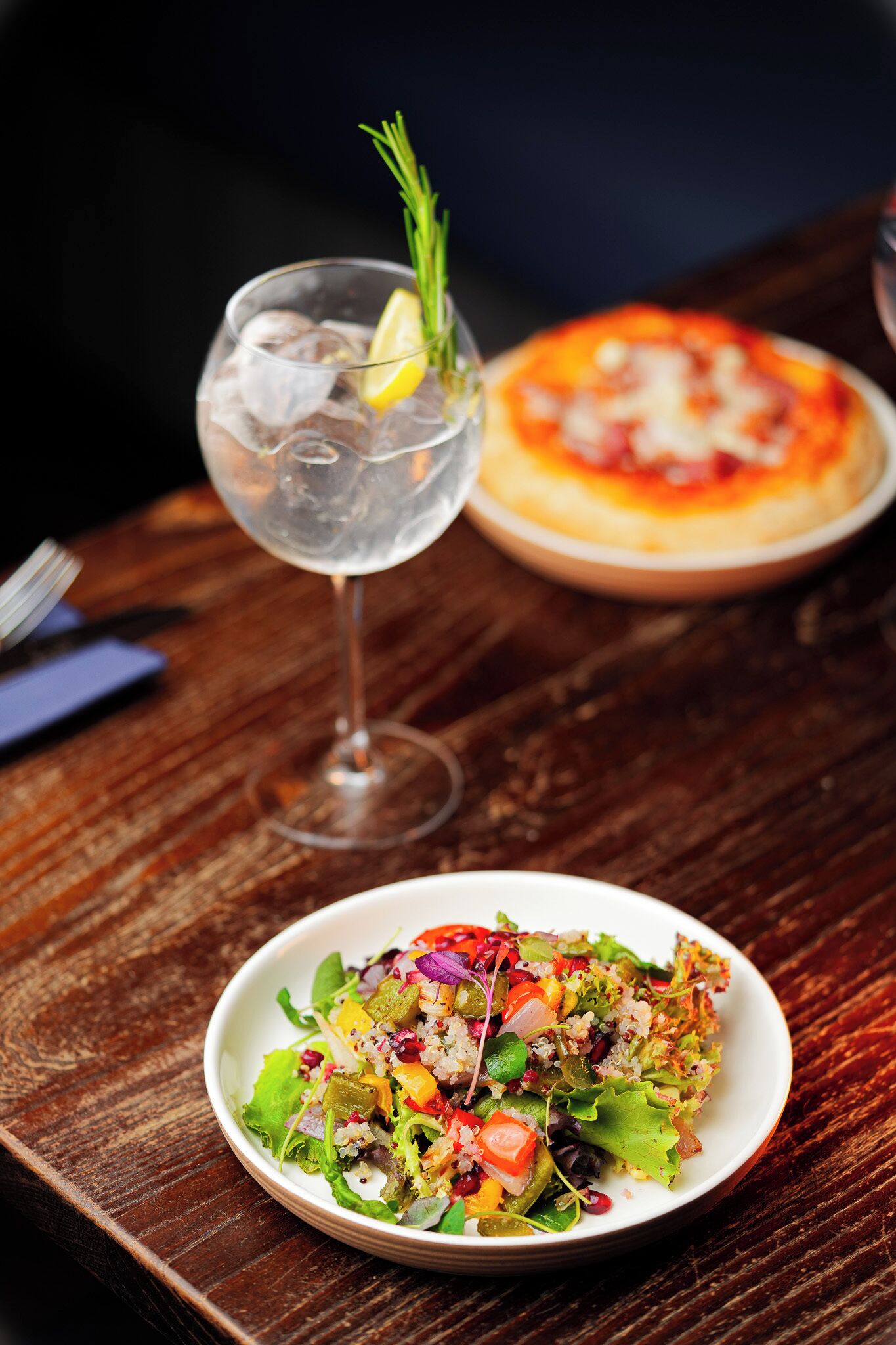Una mesa de madera con una ensalada en un plato, un vaso de agua con una rodaja de limón y una ramita de romero, y una pizza al fondo.