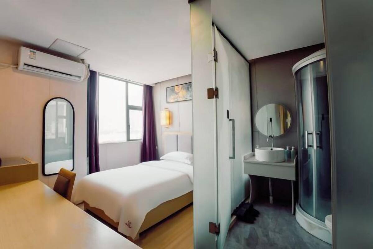 A hotel room with a bed, a desk, a mirror, an air conditioning unit, a bathroom with a sink and shower.