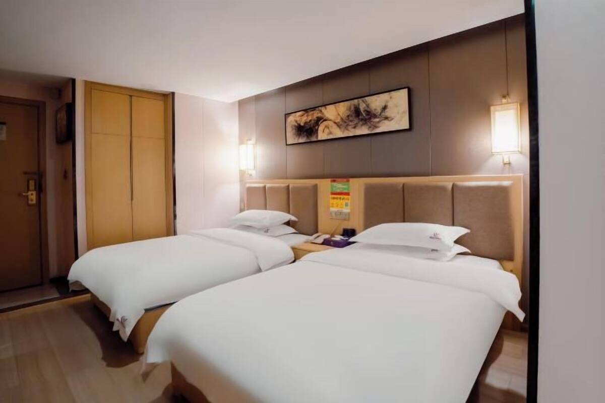 A hotel room with two beds, a wooden door, a wall-mounted artwork, and a bedside table with a lamp.
