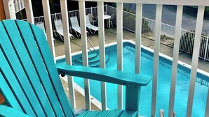 A turquoise Adirondack chair overlooking a swimming pool with lounge chairs and a white fence.