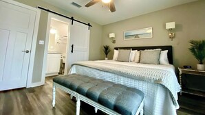 A bedroom with a bed, a bench, a ceiling fan, a door, and a nightstand with a plant.