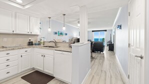 Una cocina moderna con armarios blancos, una encimera de granito y un lavavajillas empotrado. En el suelo, se observa una alfombra marrón, y al fondo, un comedor con una silla y una mesa negros.