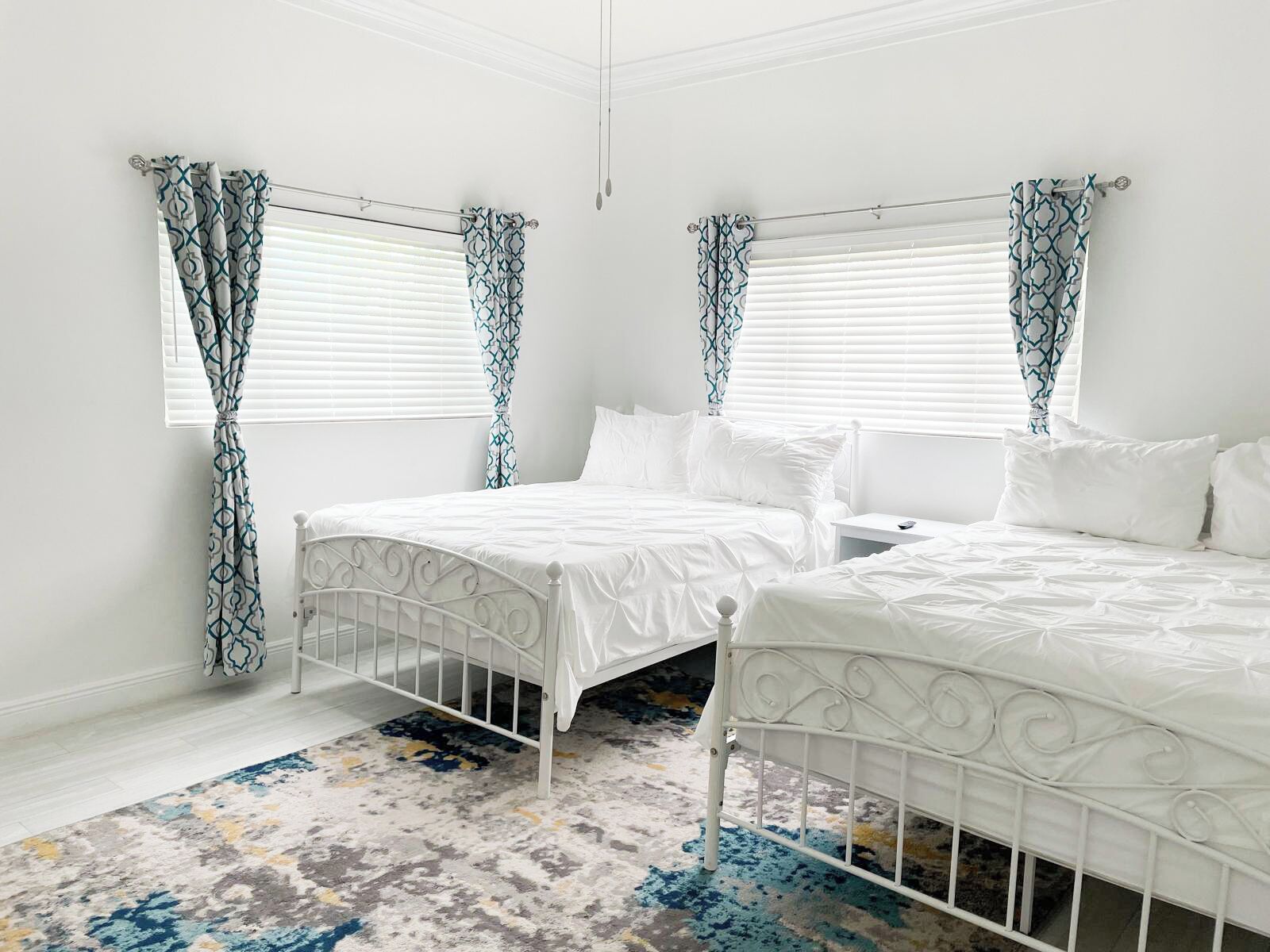 Une chambre avec deux lits, du linge de lit blanc, des rideaux à motifs et un tapis coloré.