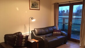 Un salon avec un canapé en cuir marron, un fauteuil plus petit, une lampe sur pied et une grande fenêtre avec des rideaux.