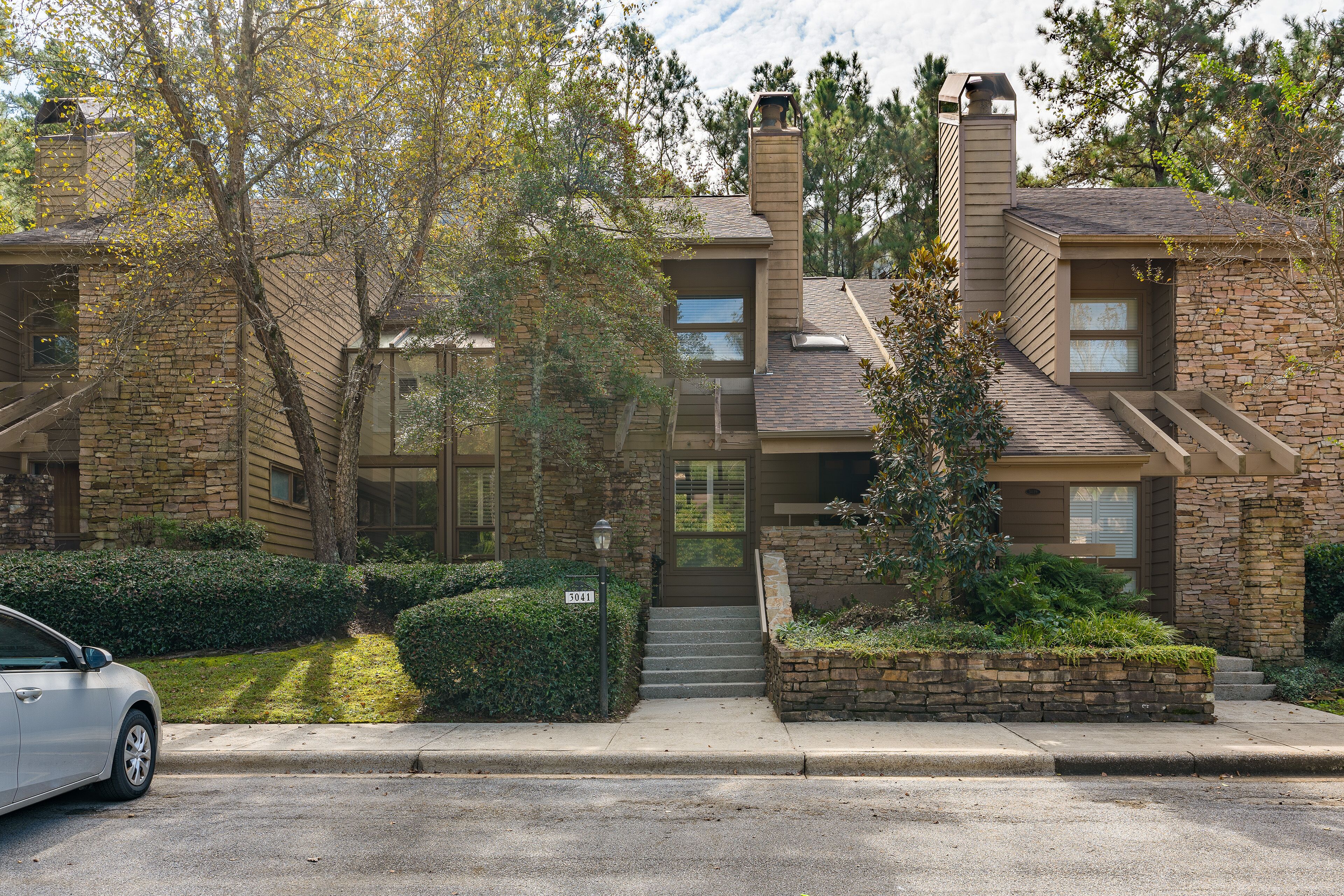 A modern two-story house with a stone facade, a chimney, and a small front yard with a car parked on the street.