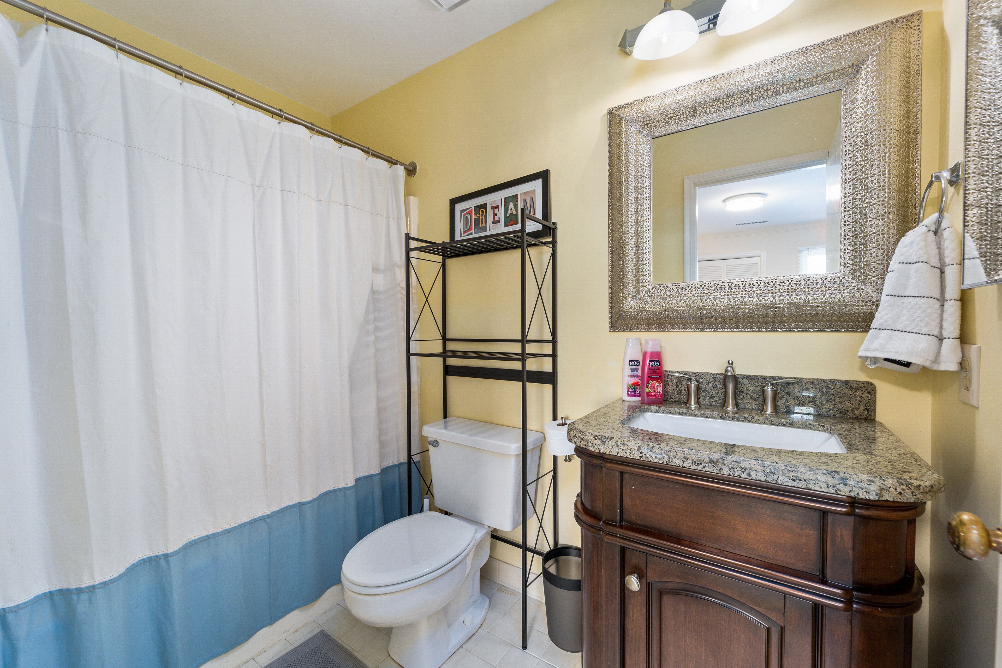 Une salle de bain avec des toilettes blanches, un rideau de douche, un lavabo avec un plan de travail en granit et un miroir doté d’un cadre décoratif.