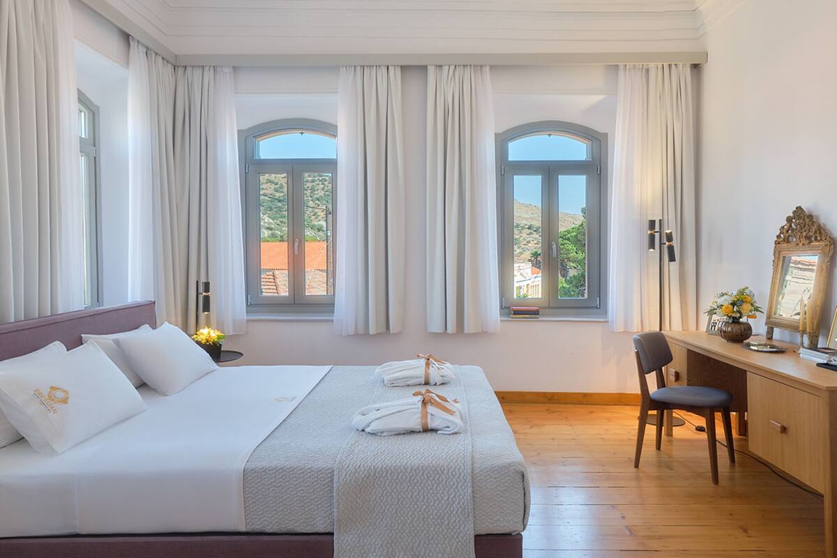 Ein geräumiges Schlafzimmer mit einem großen Bett, zwei mit Vorhängen versehenen Fenstern, einem Holztisch und einem dekorativen Stuckdekor an der Decke.
