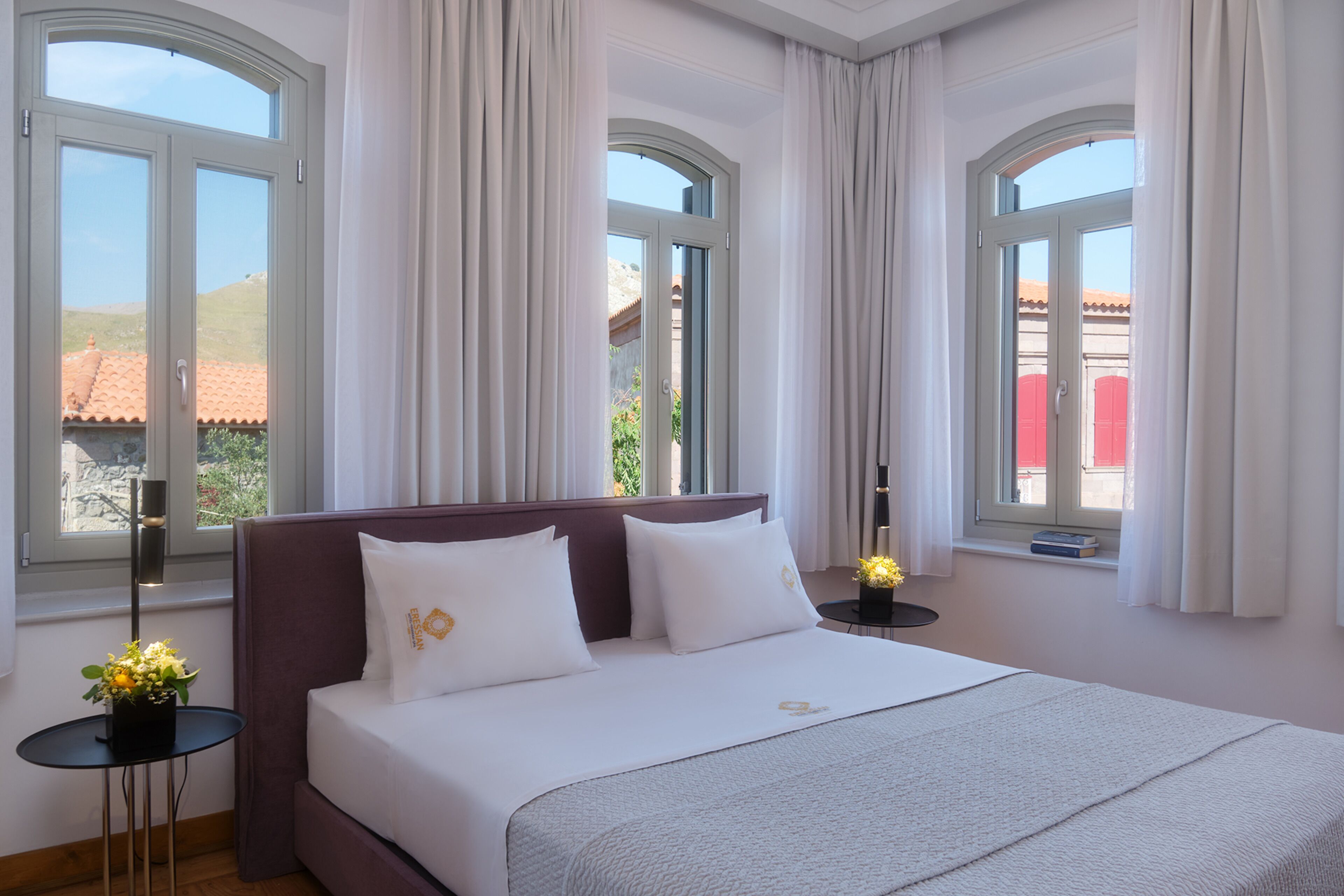 A hotel room with a large bed, two bedside tables, and a view of a red-roofed building through the windows.