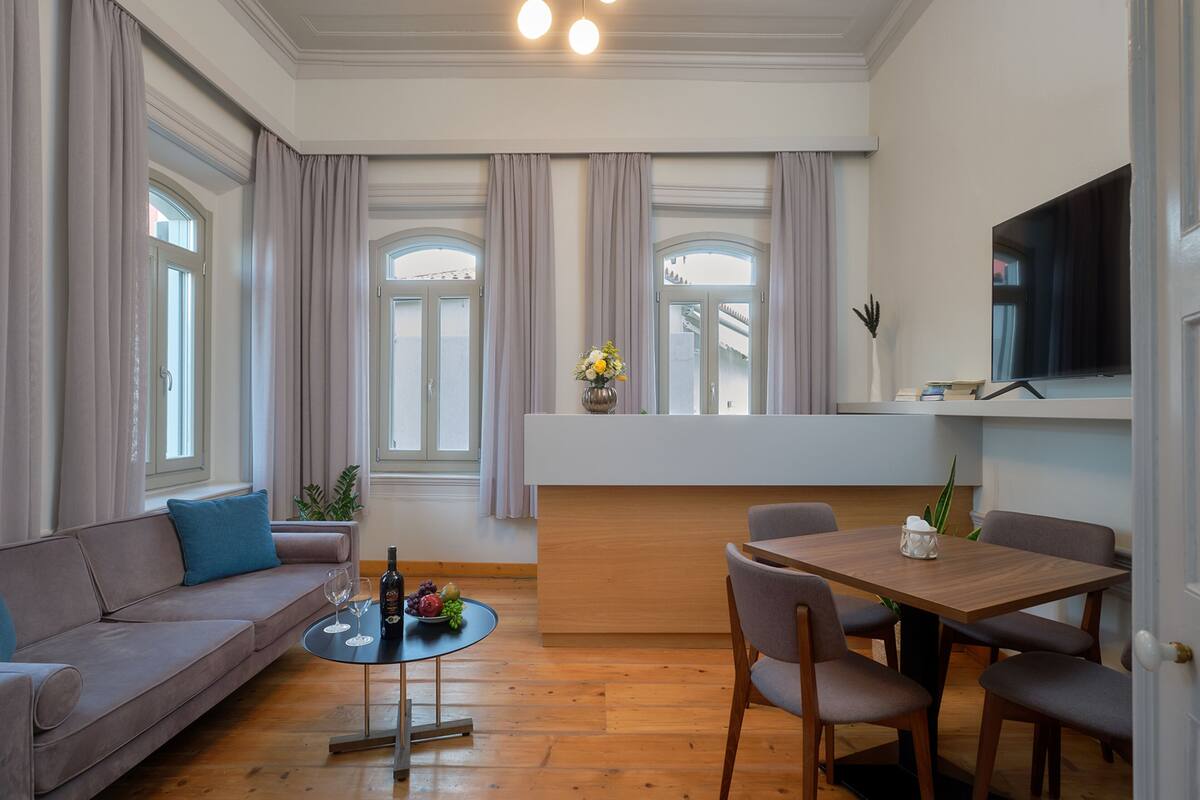 Ein modernes Wohnzimmer mit einer Couch, einem Esstisch mit Stühlen, einem Couchtisch mit einer Flasche und Obst sowie einem an der Wand montierten Fernseher.