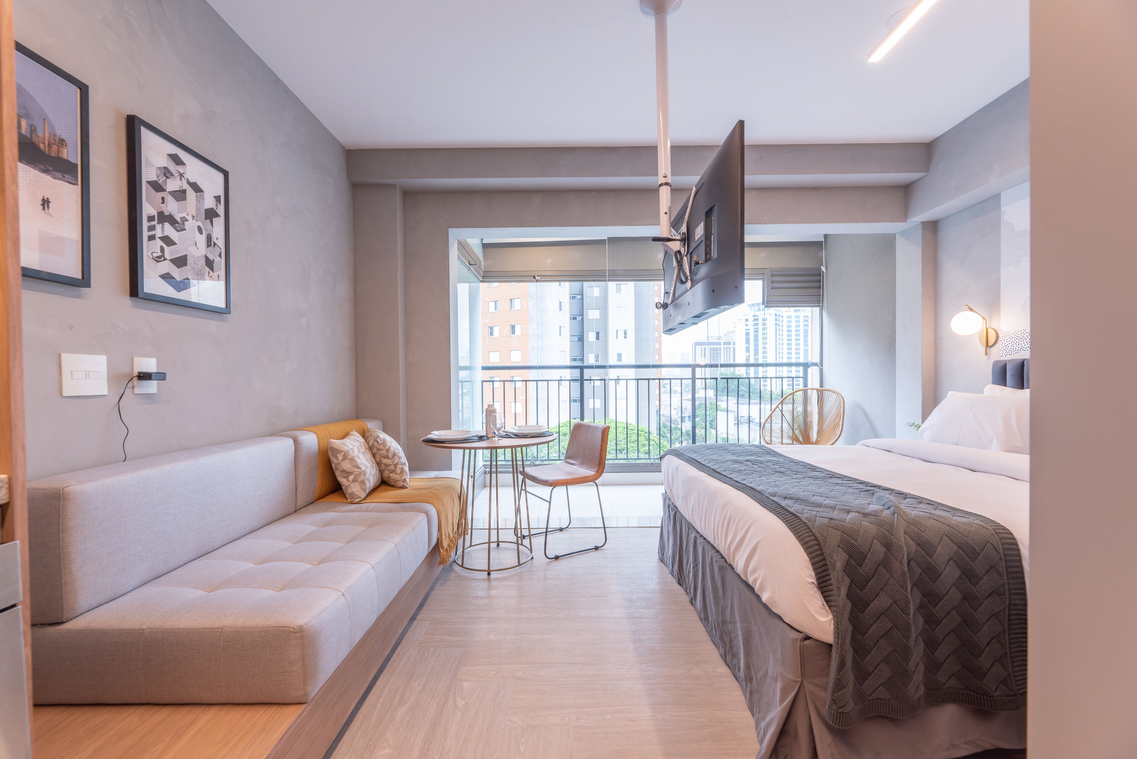 Quarto de hotel moderno com sofá, cama, mesinha com cadeira, TV e varanda com vista para edifícios.