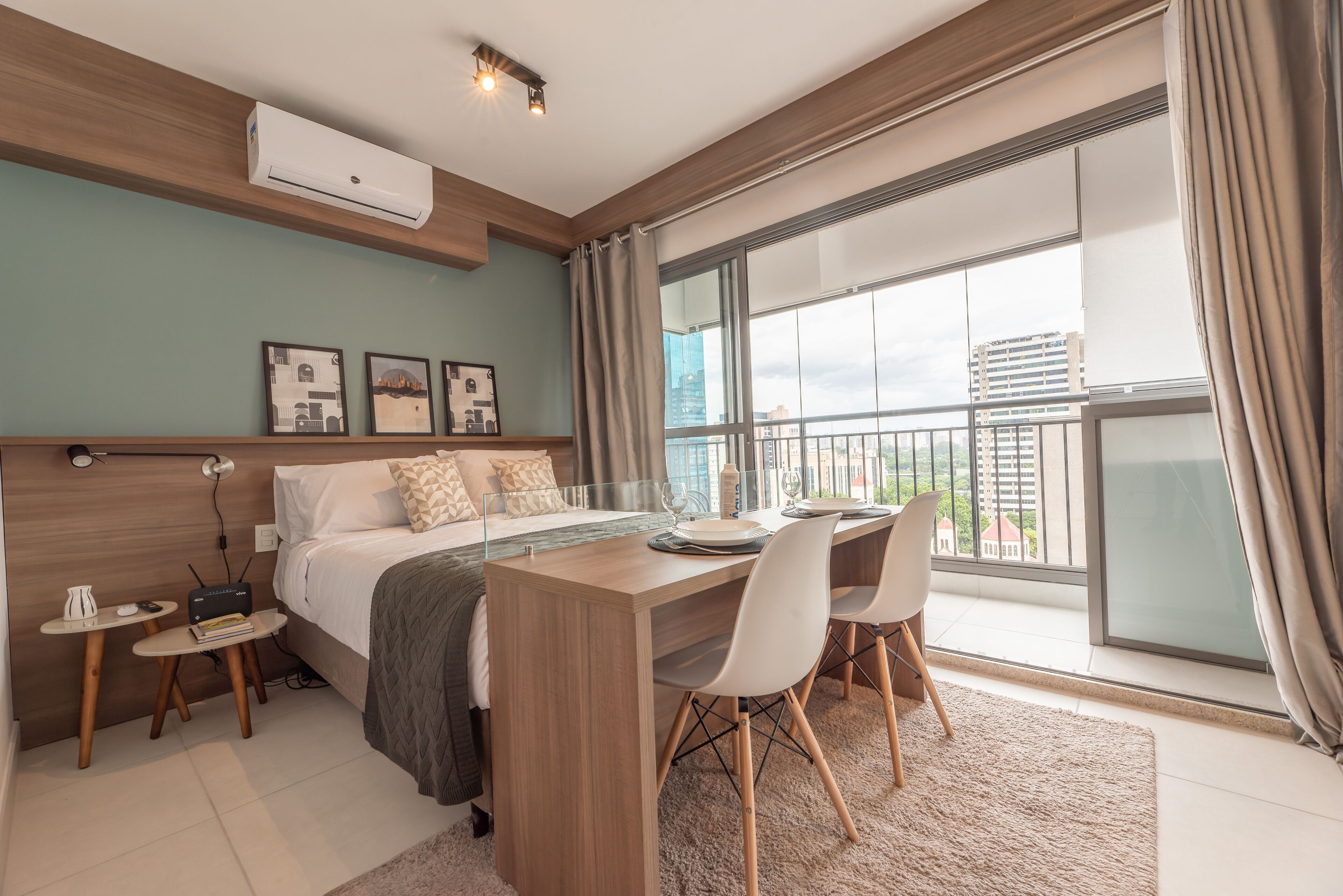 A modern bedroom with a bed, bedside tables, a desk, and chairs. There is a large window with a view of buildings.