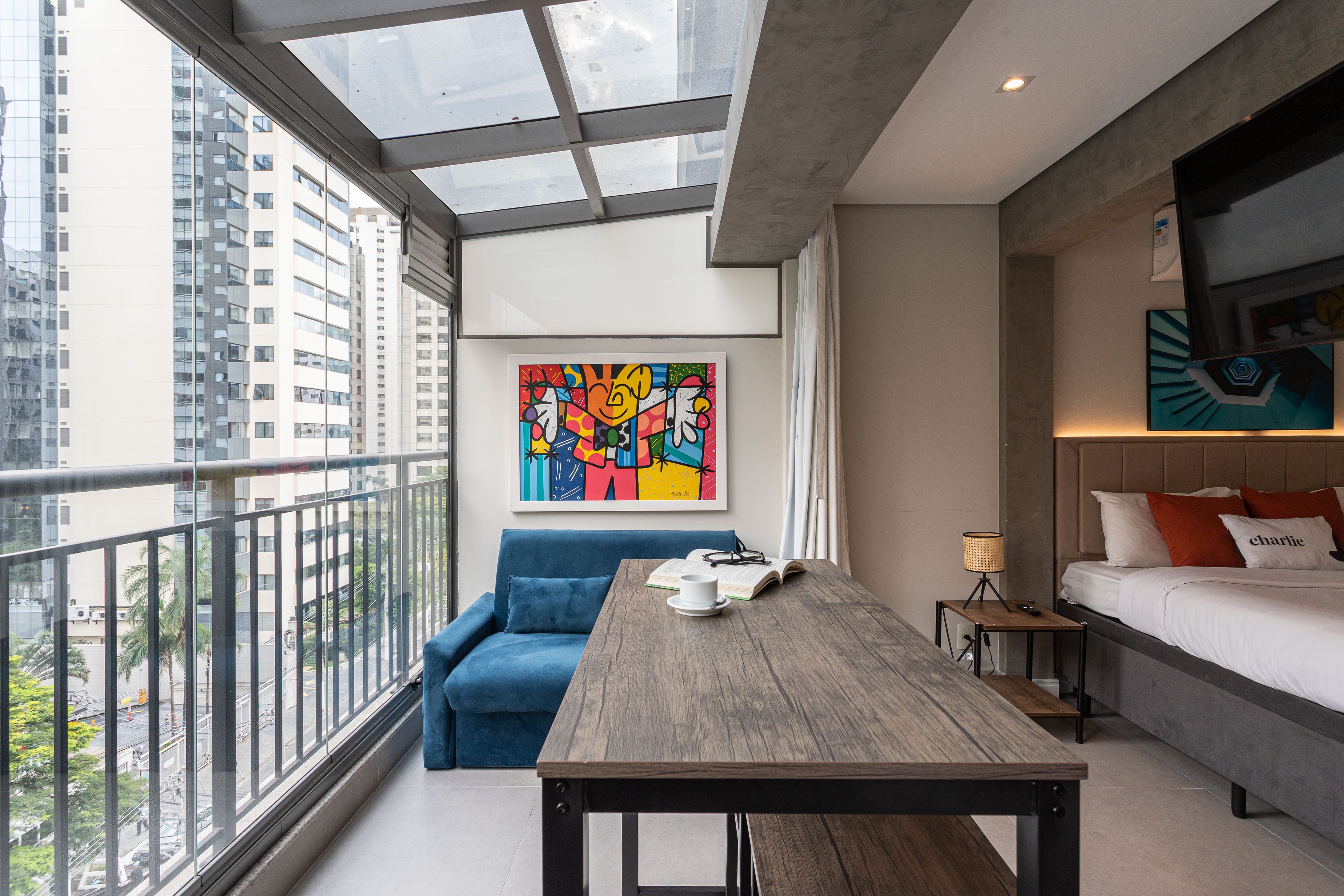 A modern hotel room with a large window, a blue sofa, a wooden table, a bed with pillows, and a colorful artwork on the wall.