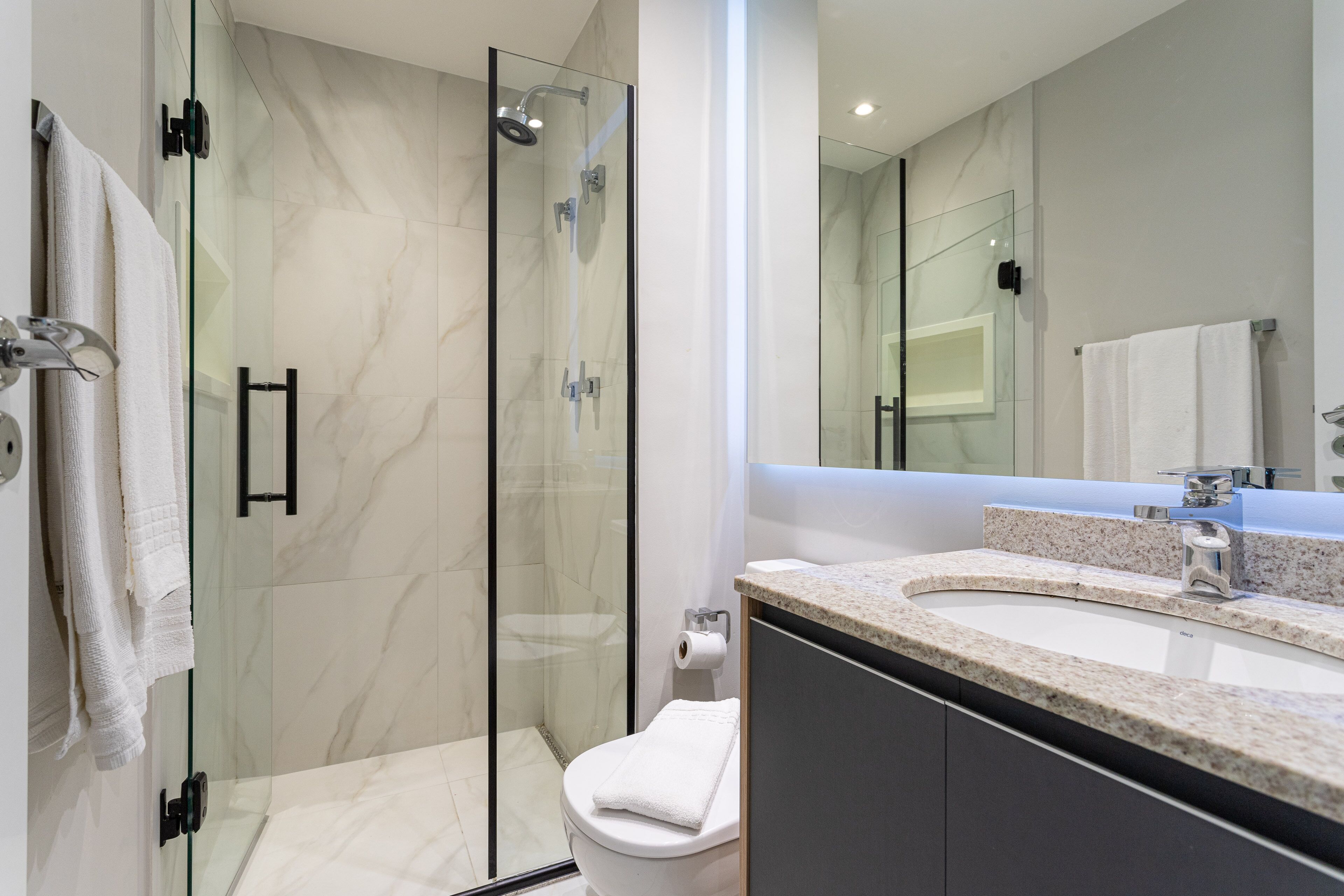 A modern bathroom with a glass-enclosed shower, a white toilet, and a granite countertop sink.