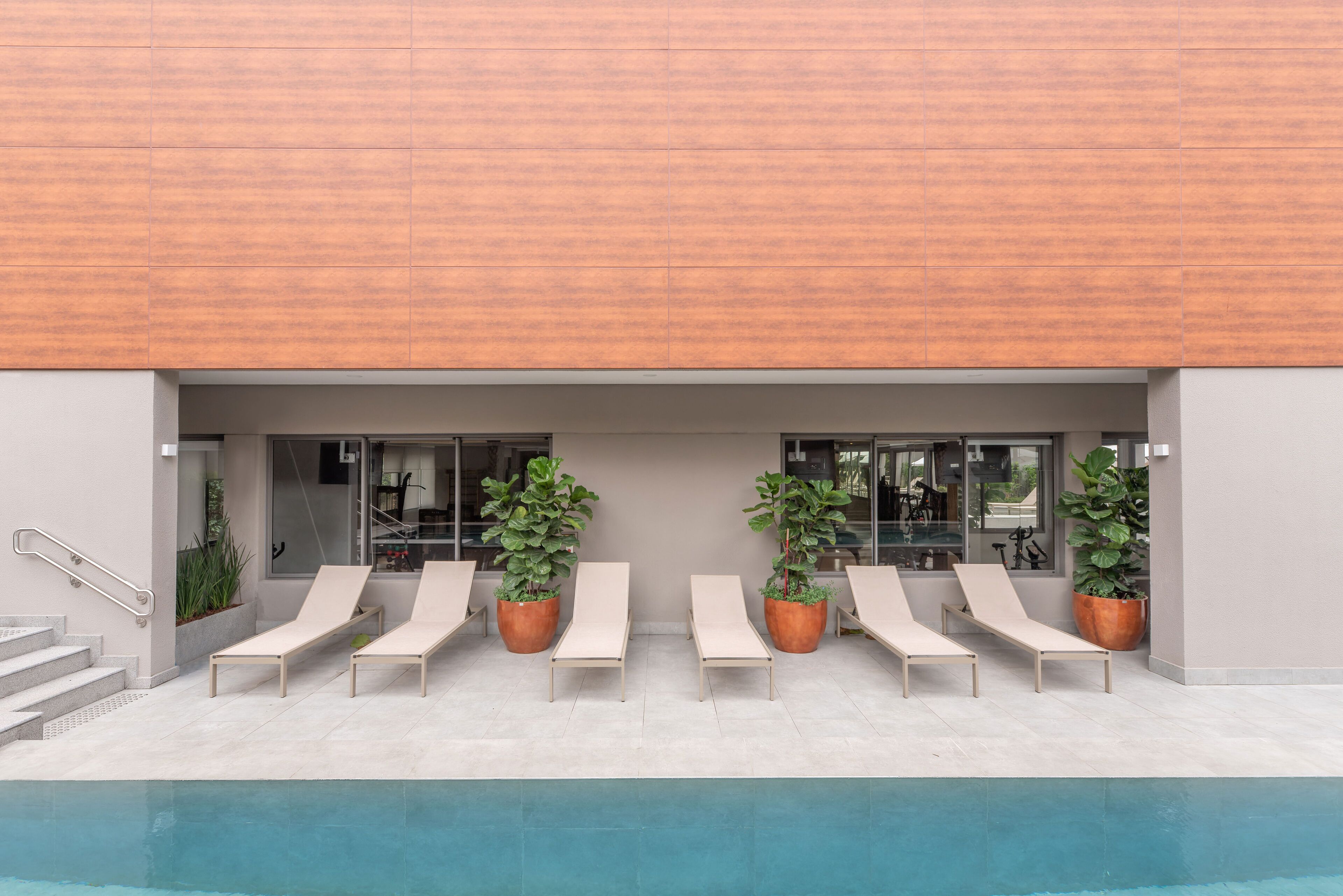Un espace aménagé au bord de la piscine, comprenant six chaises longues et des plantes en pot, un escalier et la façade d’un bâtiment moderne.