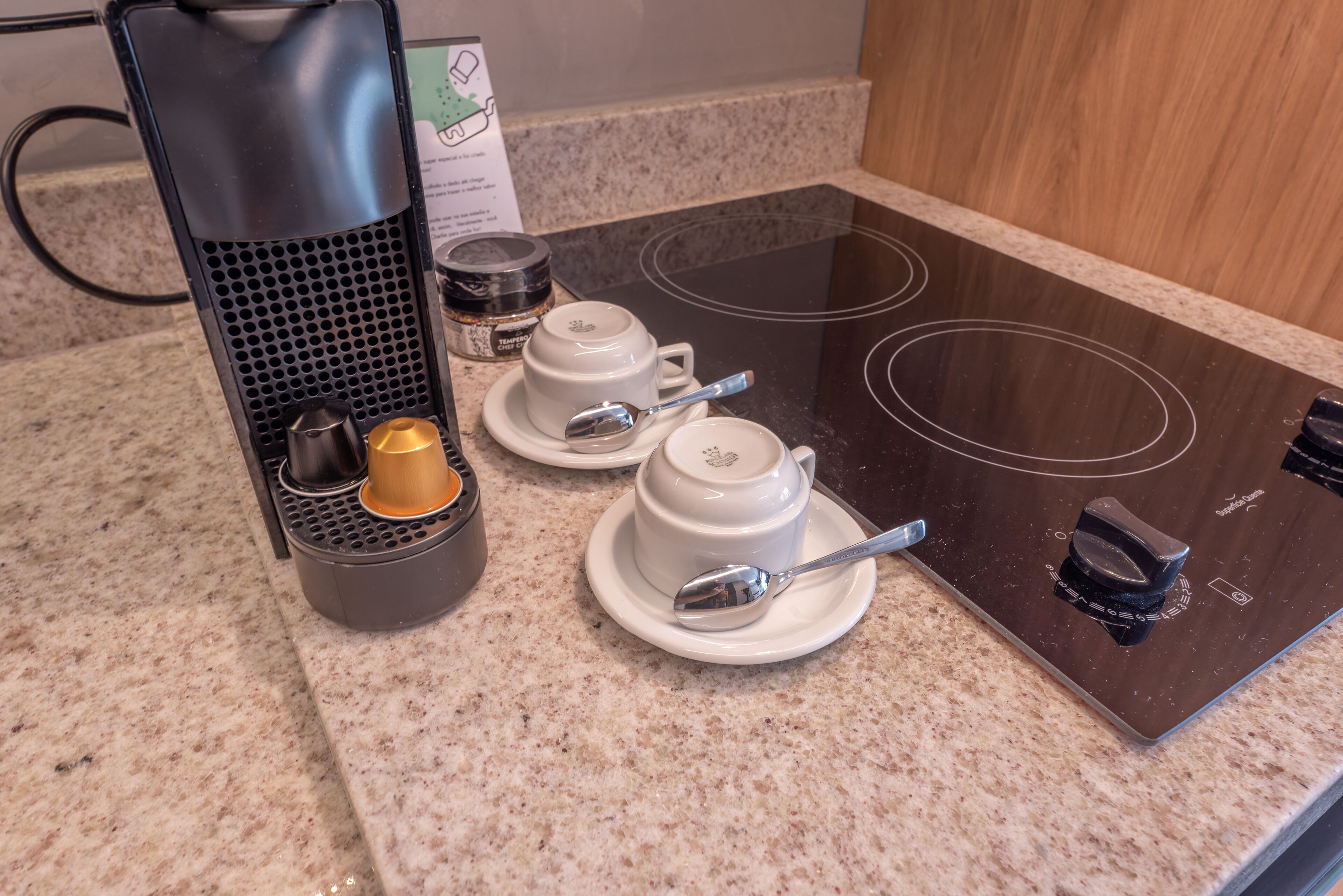 A kitchen with an induction cooktop, two white tea cups, a spoon, and a coffee maker.