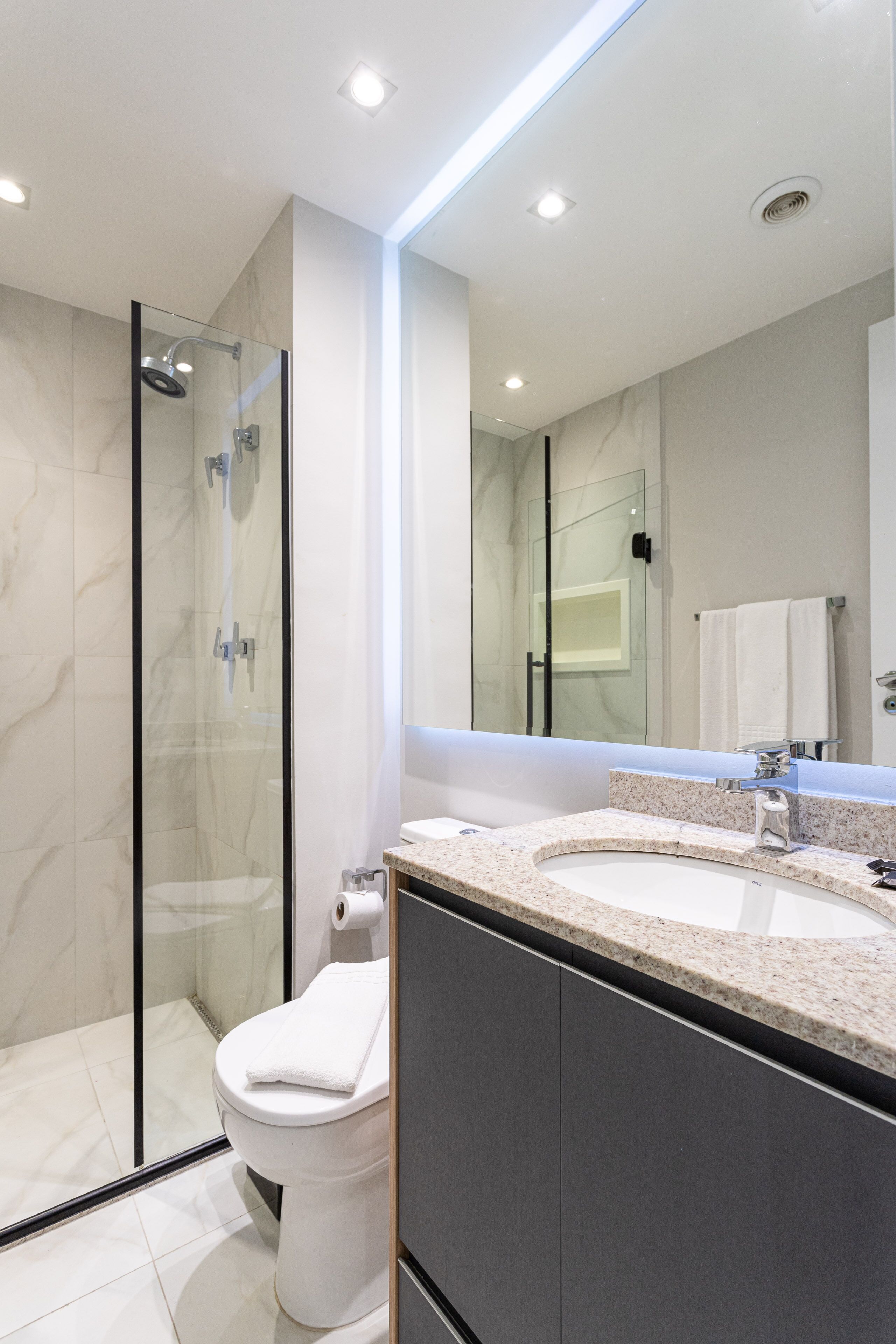 Un bany modern amb dutxa protegida per vidre, vàter blanc i un gran mirall sobre un lavabo amb encimera de granat.