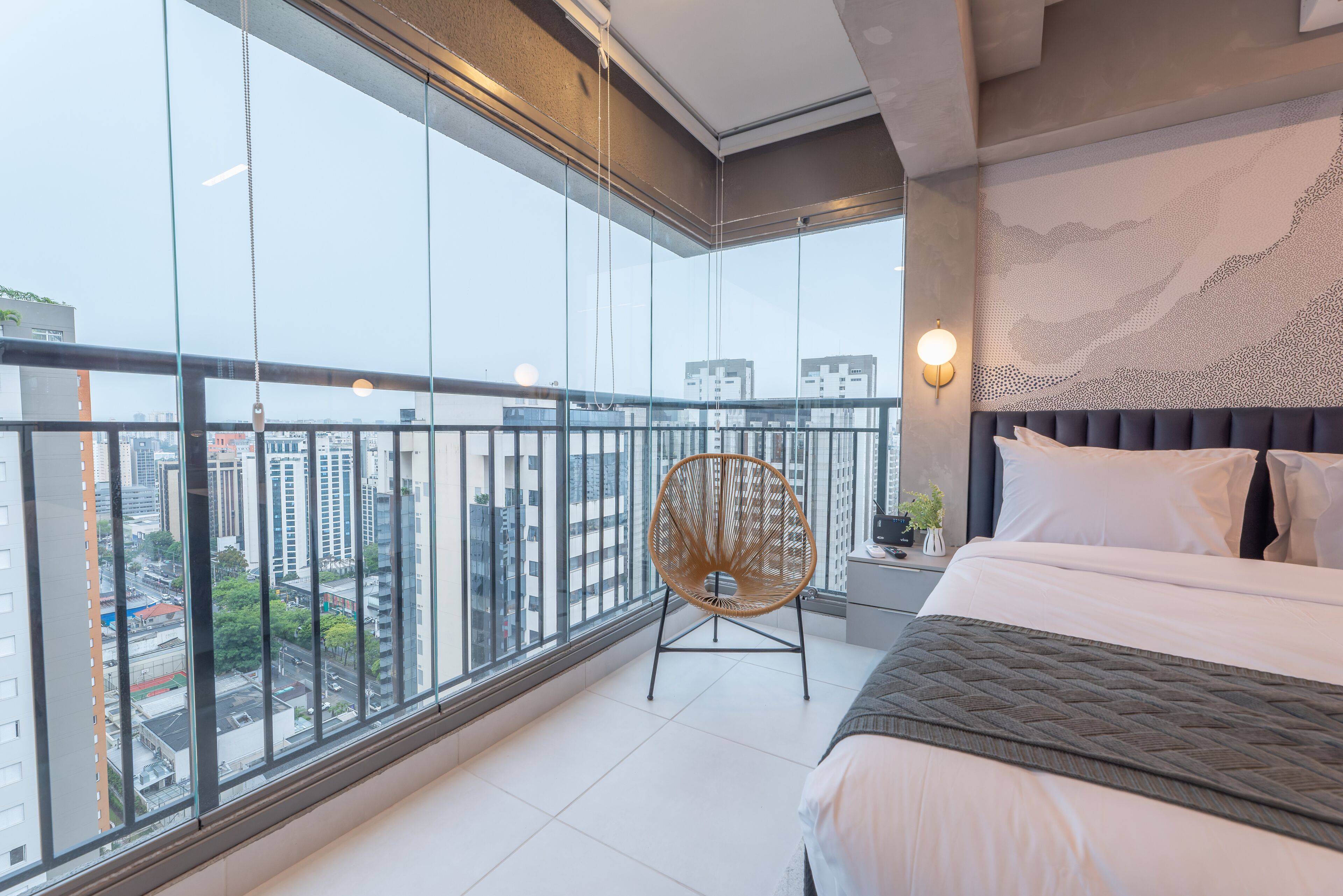 Une chambre d’hôtel moderne dotée d’un grand balcon, d’un lit recouvert d’une courtepointe matelassée, d’un fauteuil en osier et offrant une vue sur la ville.