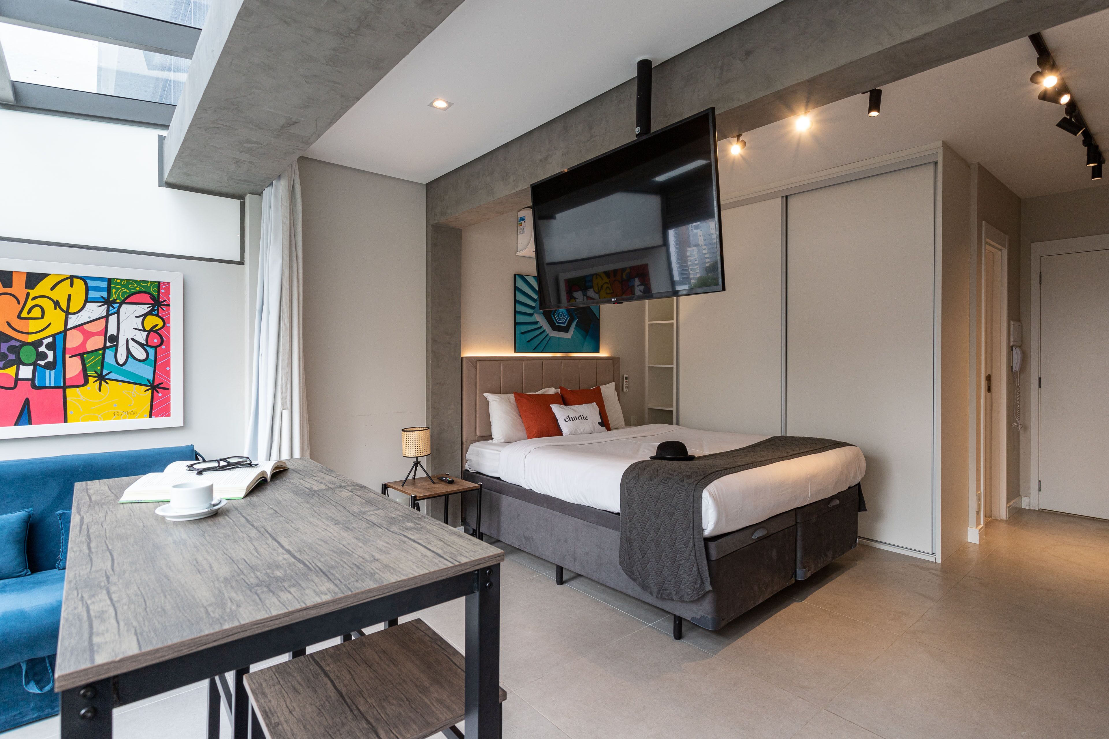 A modern hotel room with a bed, a desk, a blue sofa, and a colorful abstract painting on the wall.