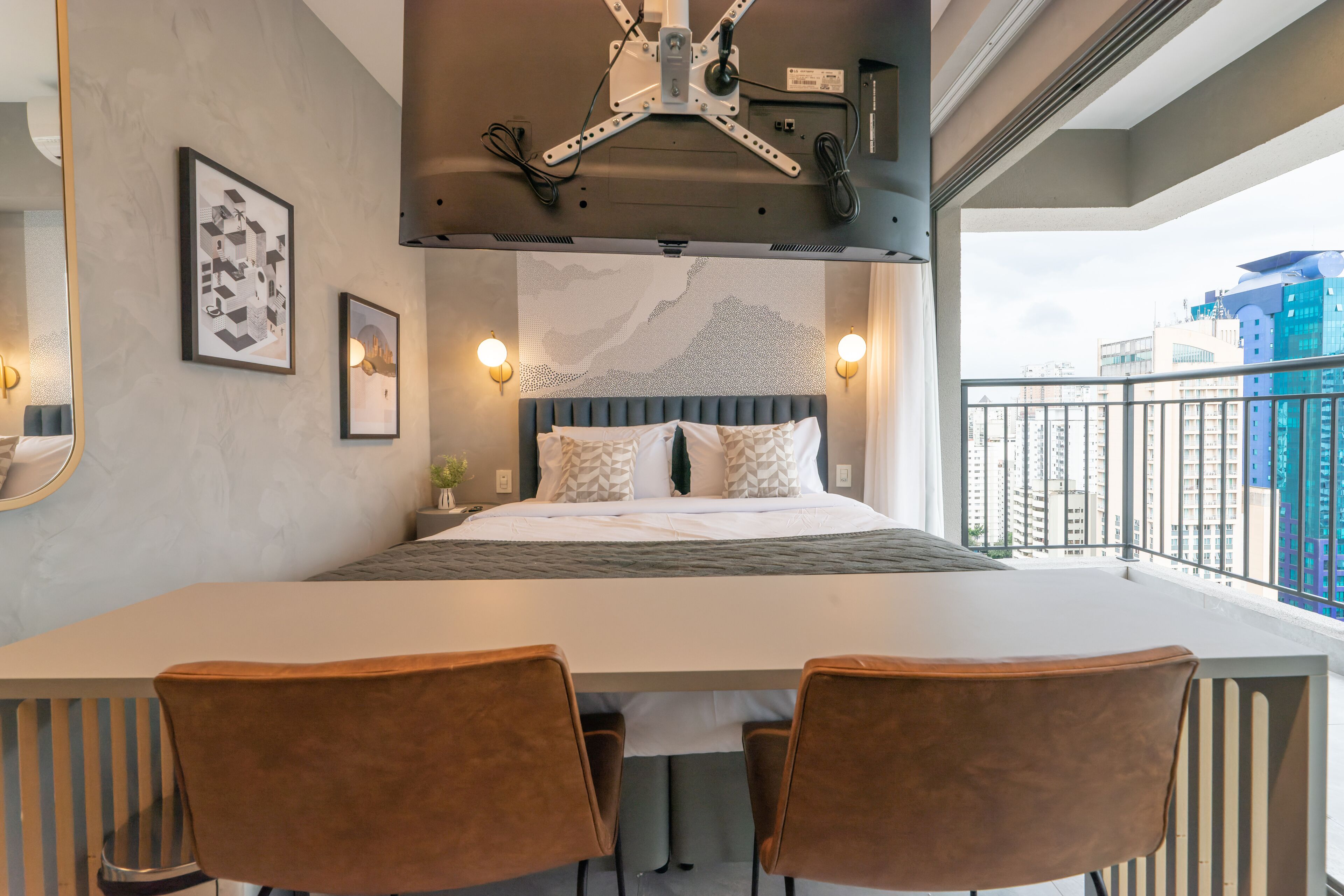 A modern hotel room with a large bed, two brown chairs, a dining table, a ceiling-mounted light fixture, and a city view.