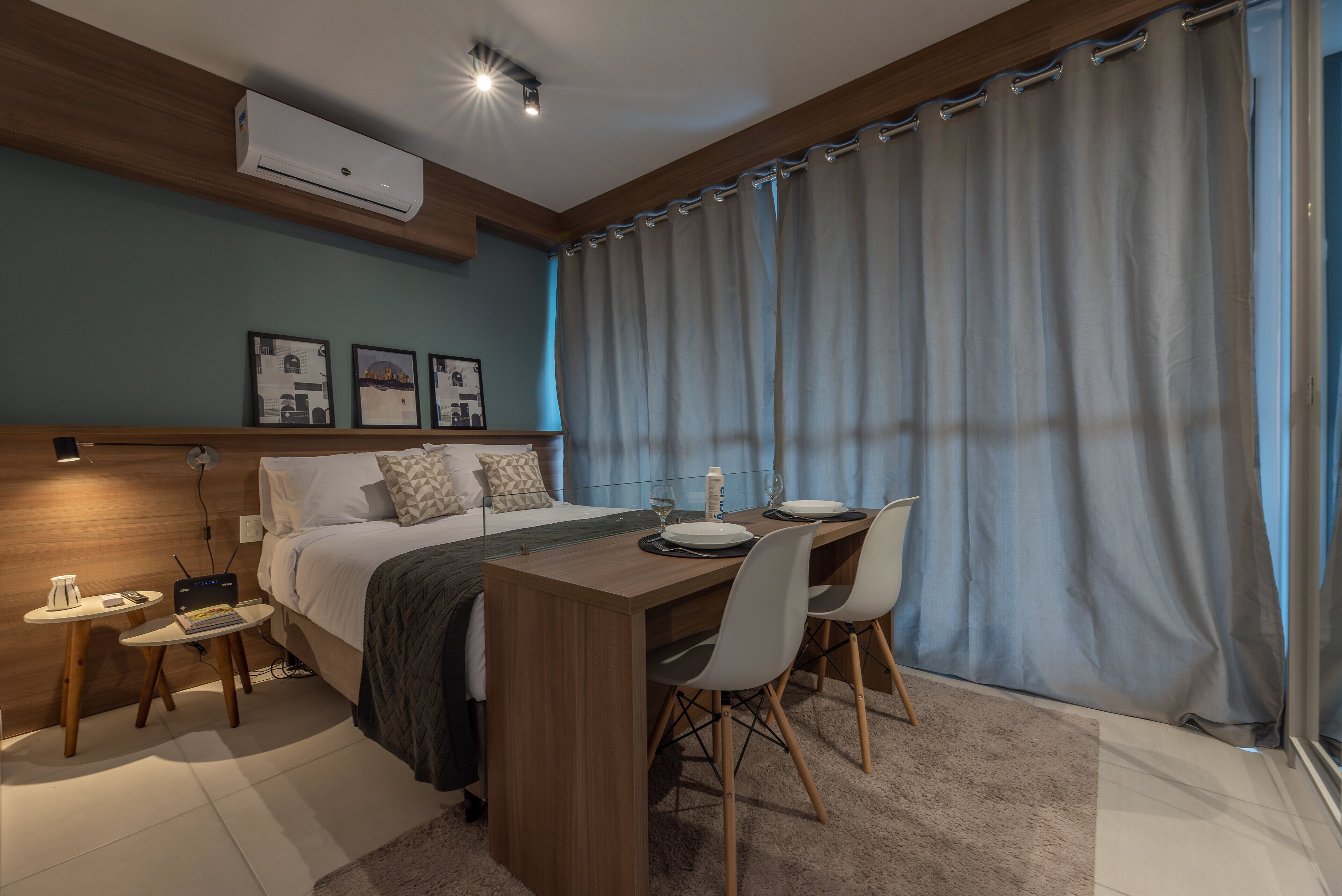 A modern bedroom with a wooden headboard, a bed with white bedding, a small bedside table with a lamp, a bedside table with a laptop, and a window with curtains.