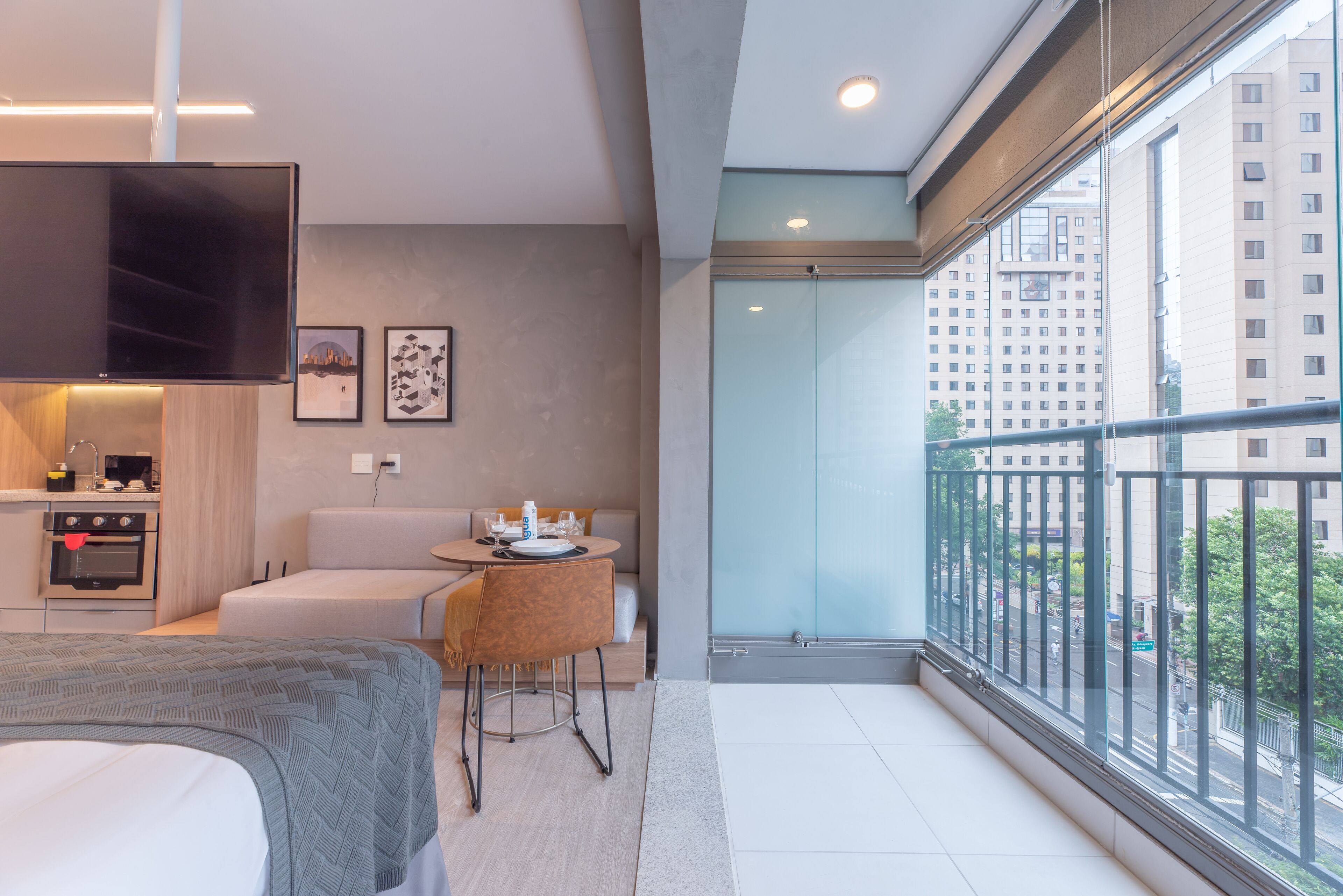 A modern hotel room with a bed, a sofa, a dining table, and a balcony with a city view.