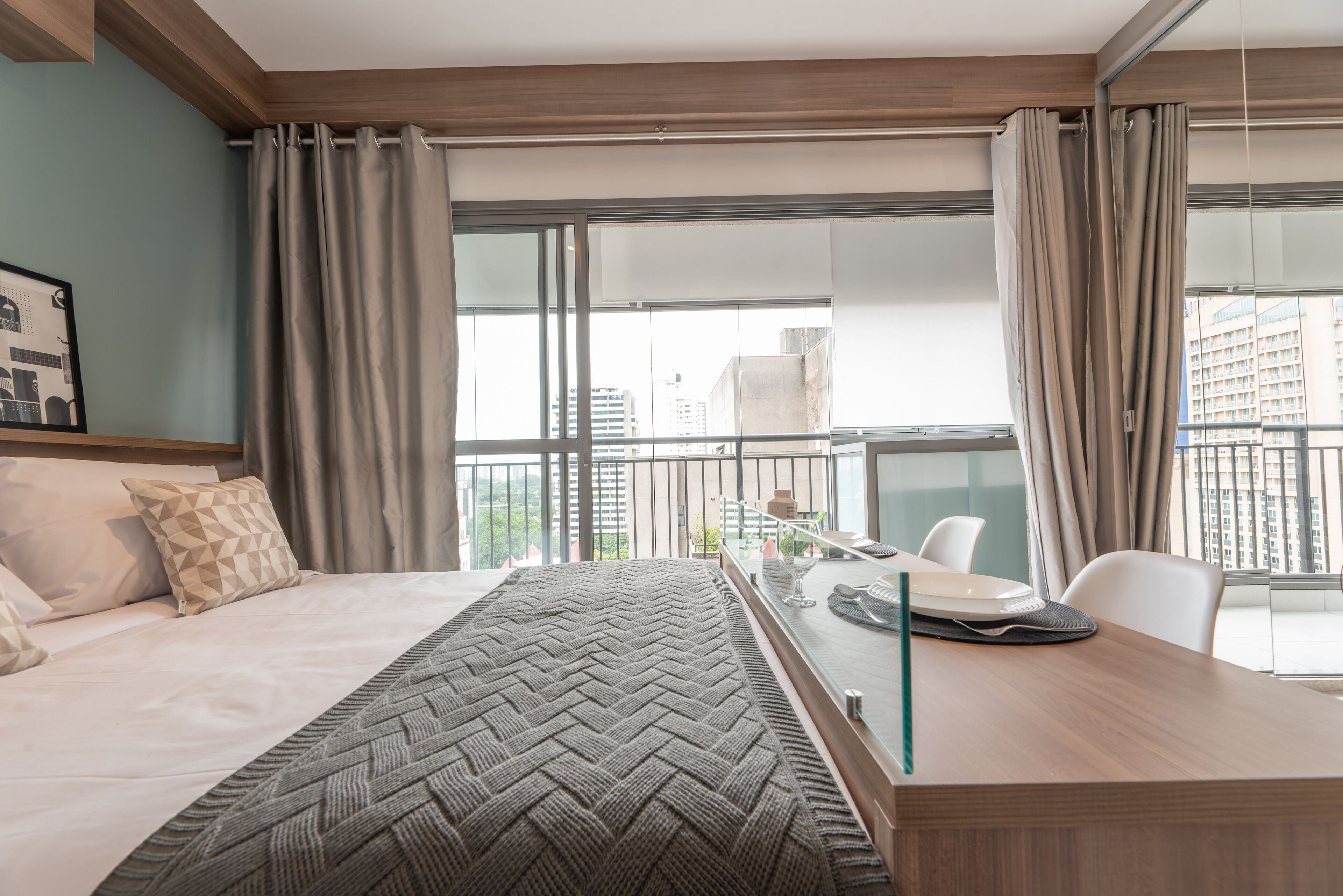 A modern hotel room with a large bed, a wooden desk, and a view of the city through large windows.