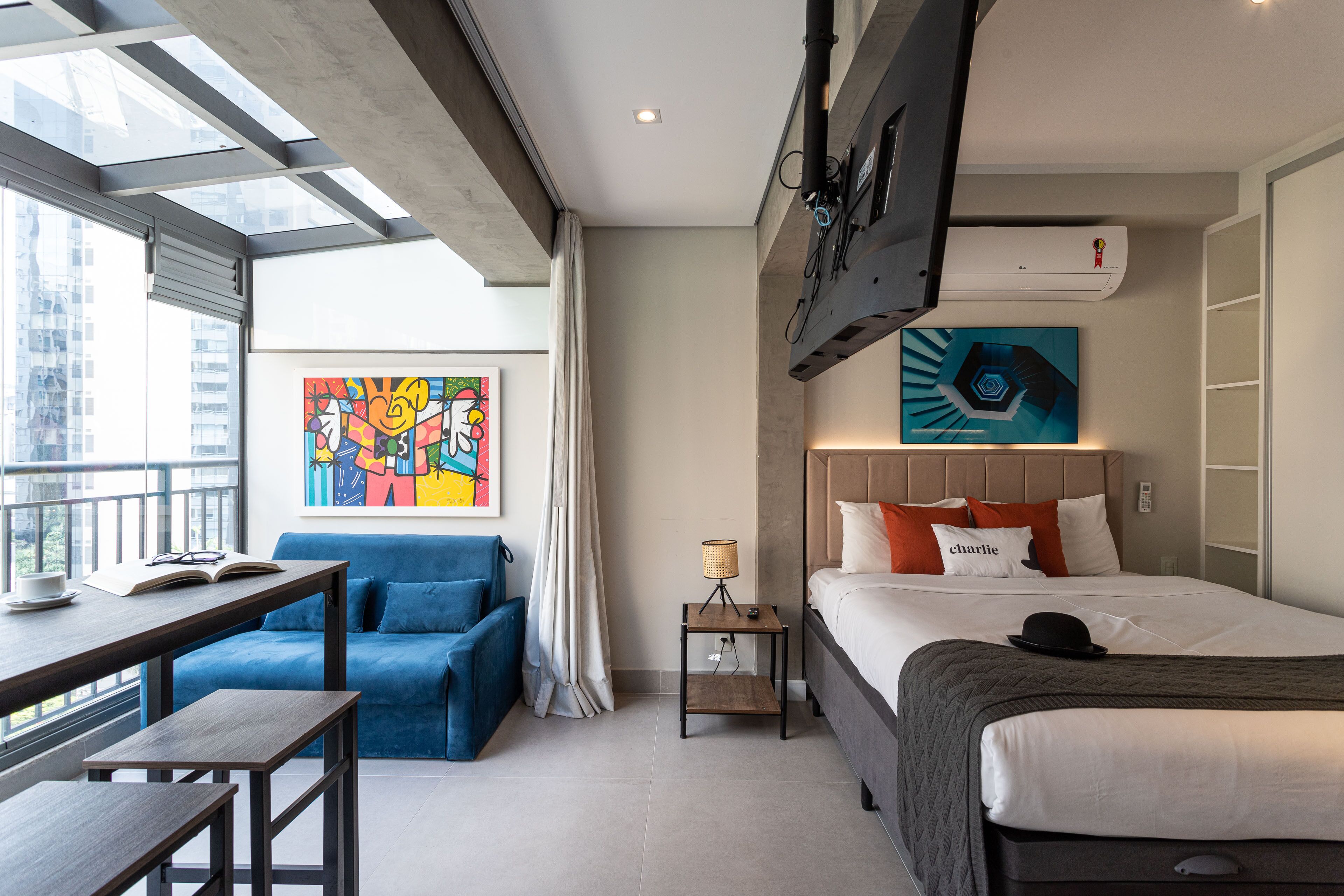 A modern hotel room with a bed, a desk, a blue sofa, a small table, a lamp, and a painting on the wall.