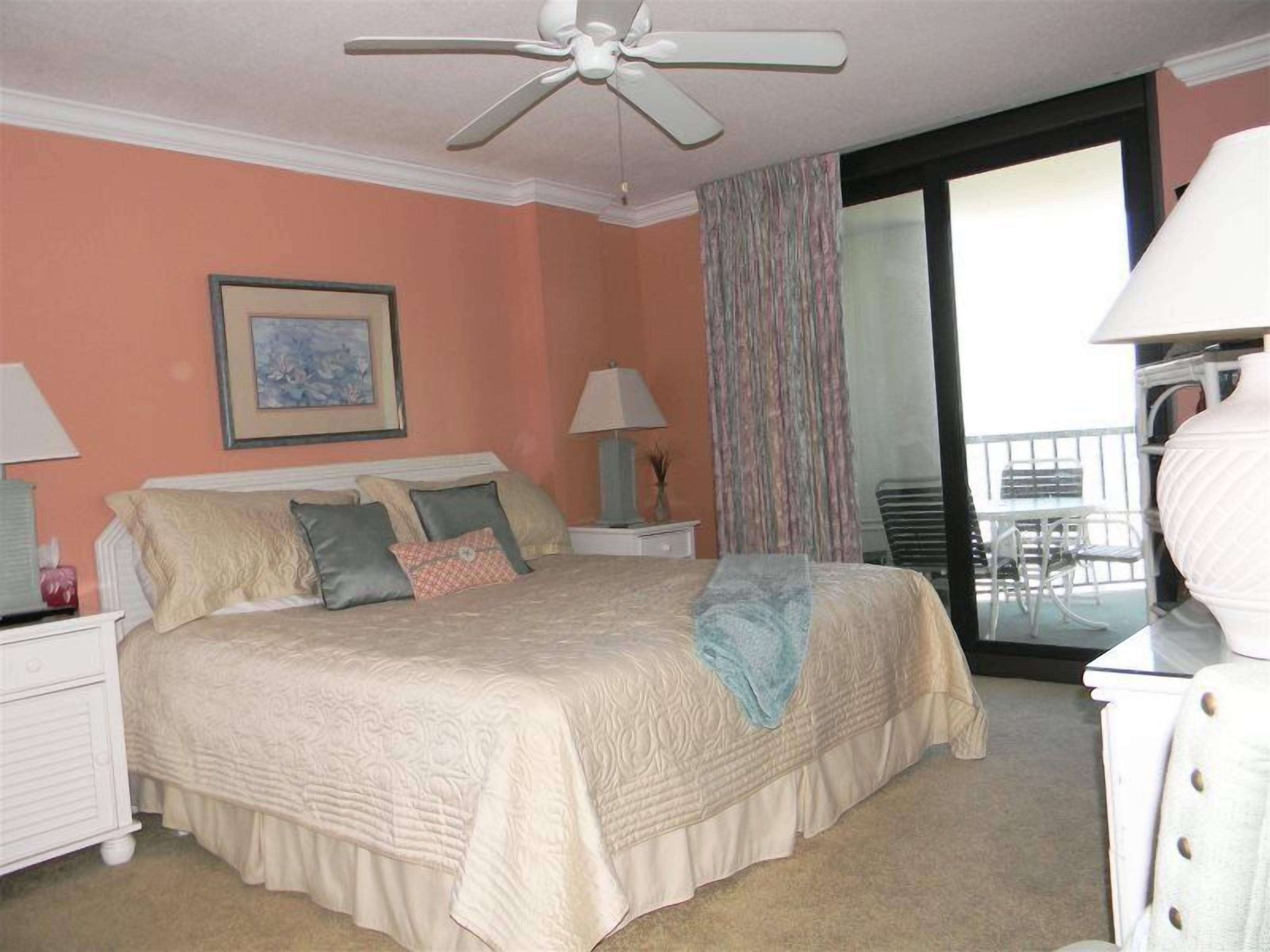 A bedroom with a bed, nightstands, a ceiling fan, a painting, a window with curtains, and a balcony with chairs.